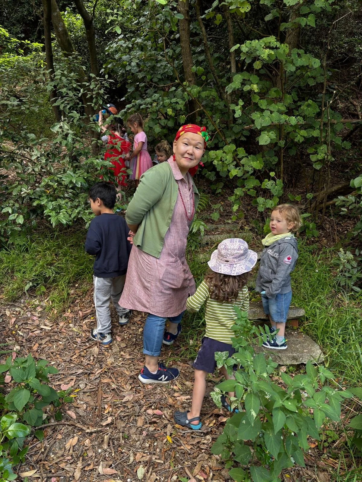 A woman and several children on a nature trail surrounded by green trees and bushes. The woman is wearing a pink dress, green cardigan, and red headscarf, and the children are dressed casually, some wearing hats. The group appears to be exploring or 