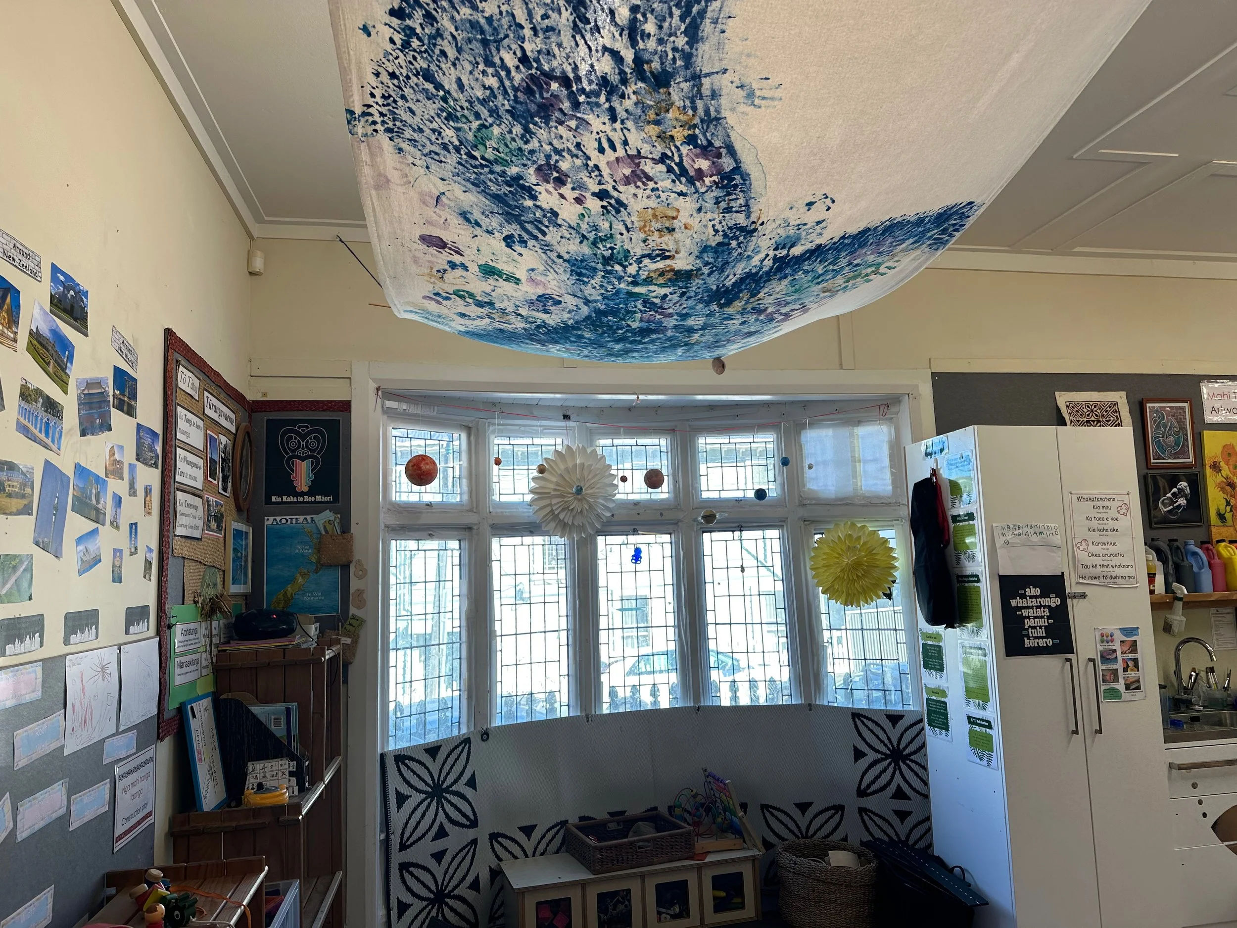 A classroom with colorful artwork on the walls, a large blue and white fabric ceiling decoration, and hanging paper decorations, including the solar system, near the window.