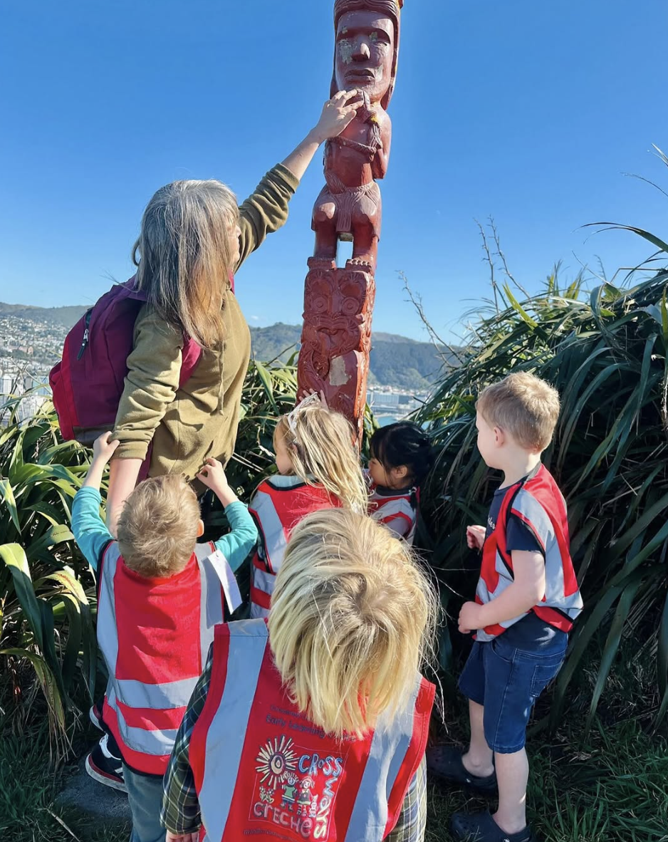 A group of young children wearing red safety vests with the word 'CROSS' and a logo are gathered around a woman, likely a teacher or guide, who is touching a tall, carved wooden totem pole with a face at the top and various designs, outdoors during d