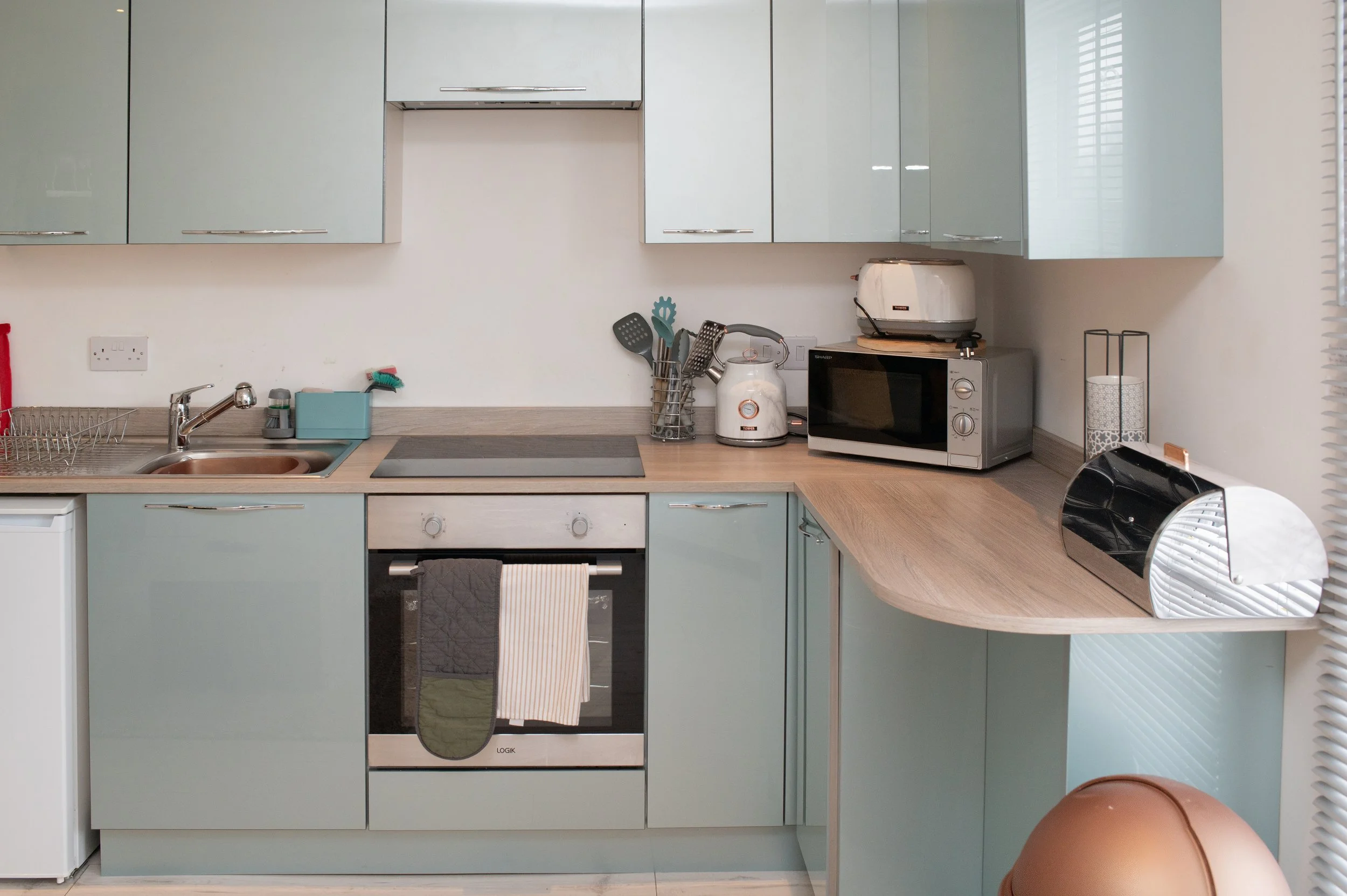 Accommodation Alsager Modern kitchen with light blue cabinets, beige countertops, and a wooden breakfast bar. Contains a microwave, toaster, utensil holder, oven with towels hanging, and a small refrigerator.
