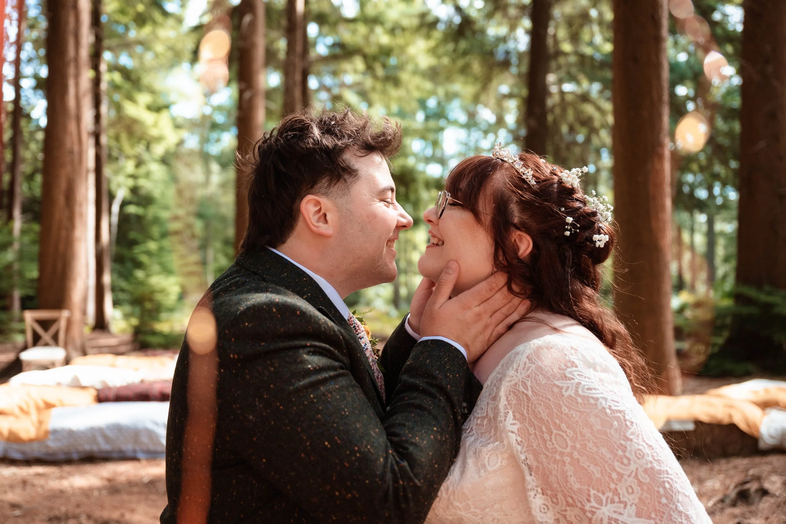 A man and woman in wedding attire sharing an intimate moment in a forest, with chairs and blankets in the background.