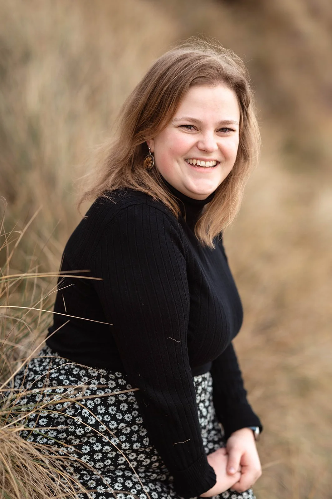 A woman with light brown hair, wearing a black long-sleeve top and floral skirt, sitting outdoors among tall grass, smiling at the camera.