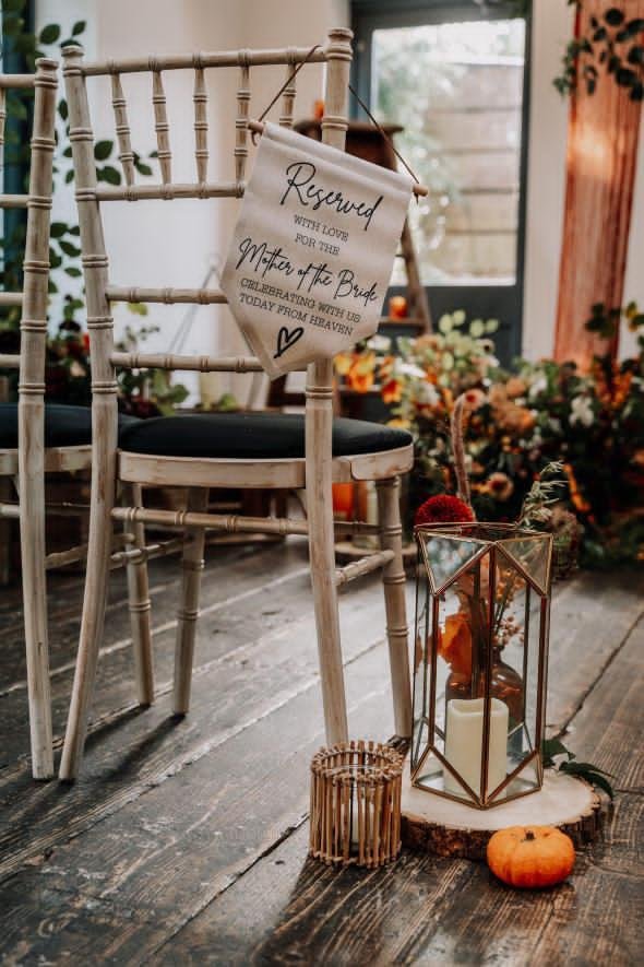 Vintage wedding seating card display with a 'Reserved' sign hanging on a white wooden chair, surrounded by autumnal decorations including a glass lantern with candles and flowers on a wooden slab, on a rustic wooden floor.