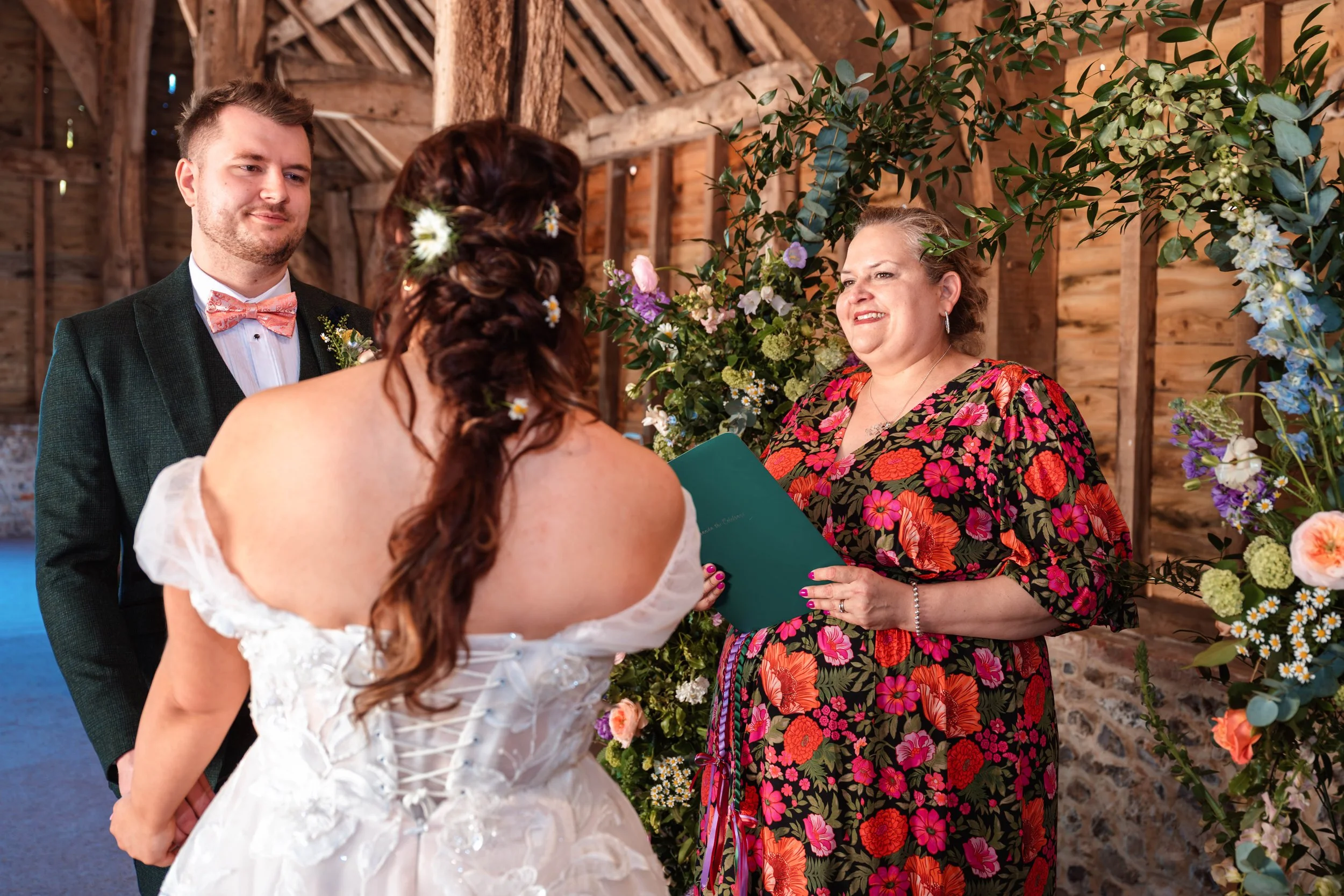 Bride and groom standing before officiant during wedding ceremony in rustic wooden setting with floral decorations.