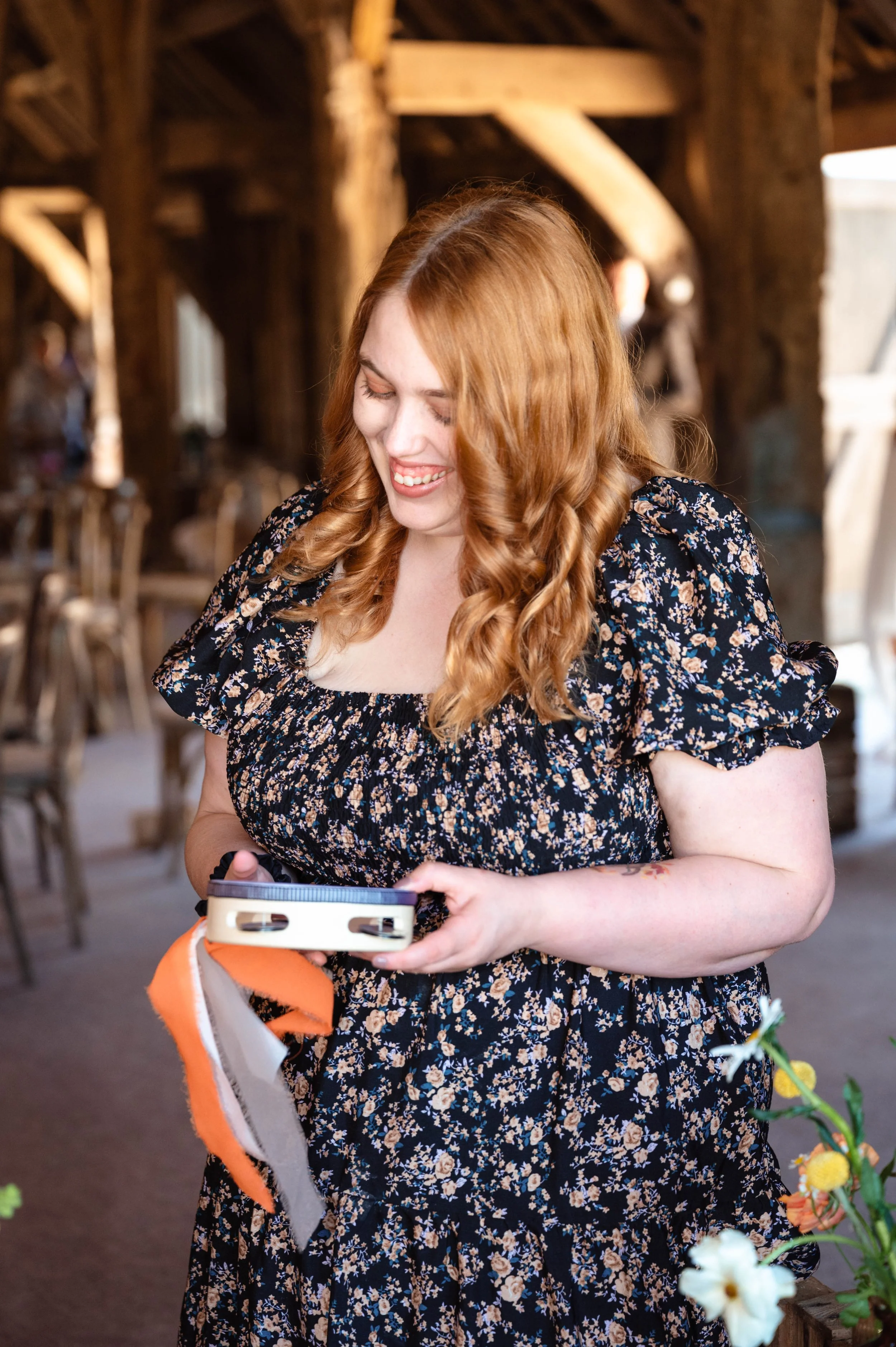A woman with long red hair in a floral dress, smiling and looking down at a label maker in her hands, inside a rustic wooden building with chairs and flowers around.