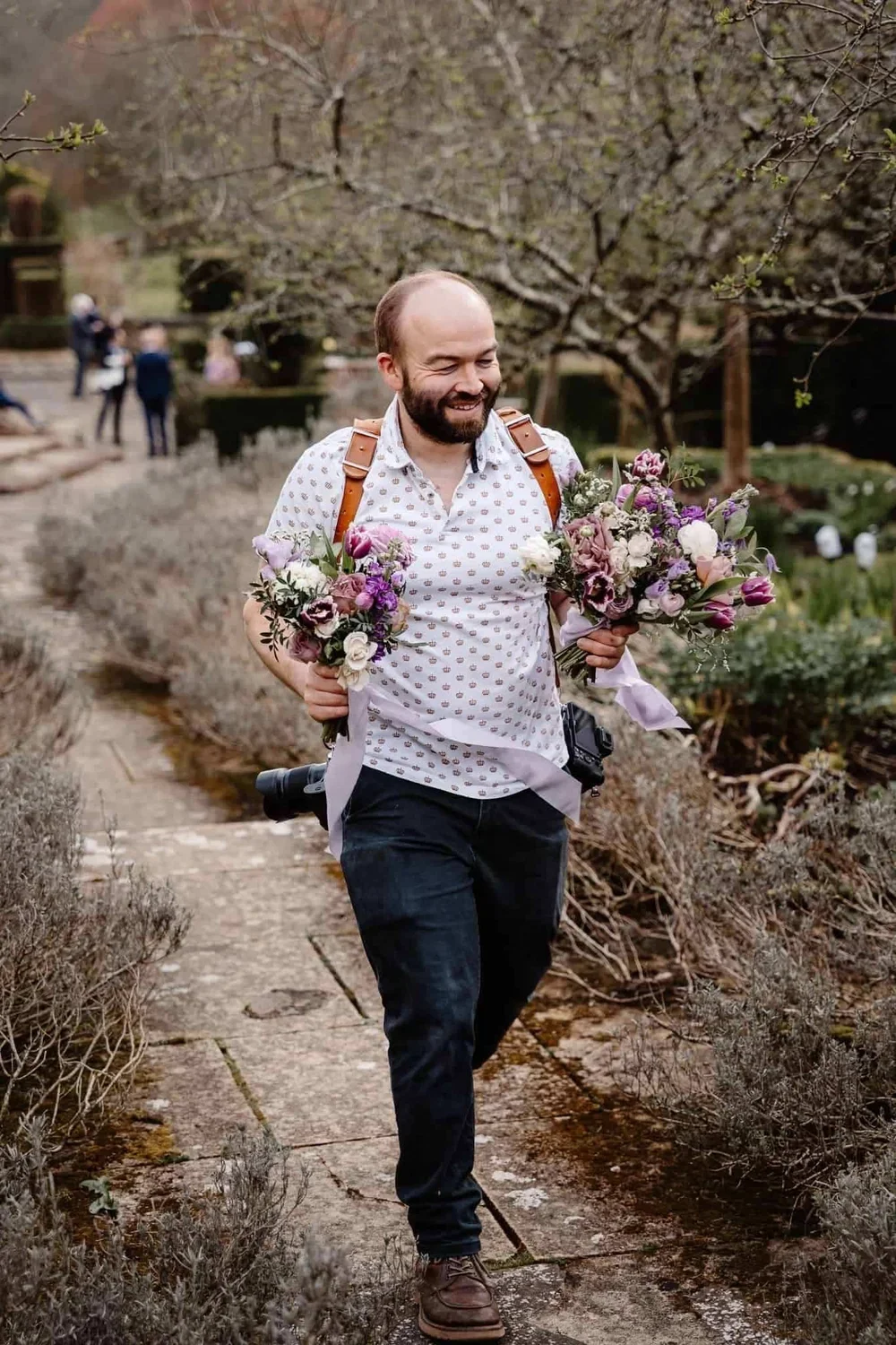 A man with a beard smiling and carrying two bouquets of flowers in a garden.
