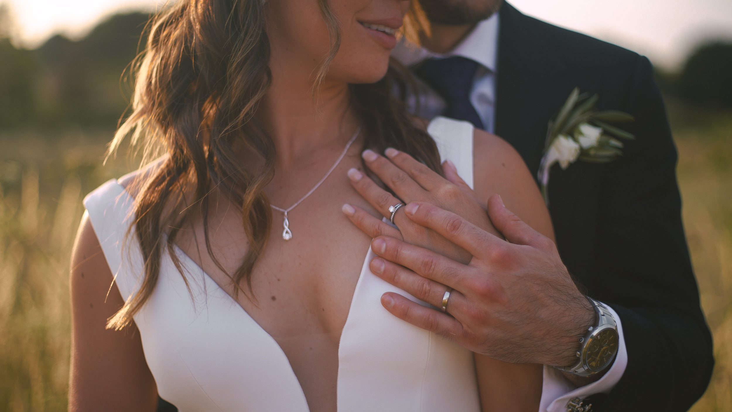 A close-up of a newly married couple, with the groom gently holding the bride's shoulder, both wearing wedding rings, in an outdoor setting at sunset.