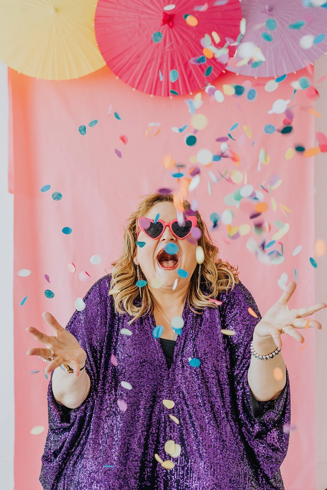 Woman wearing purple sequined top, pink heart-shaped sunglasses, and earrings, throwing colorful confetti at a celebration with pink balloons and streamers in the background.