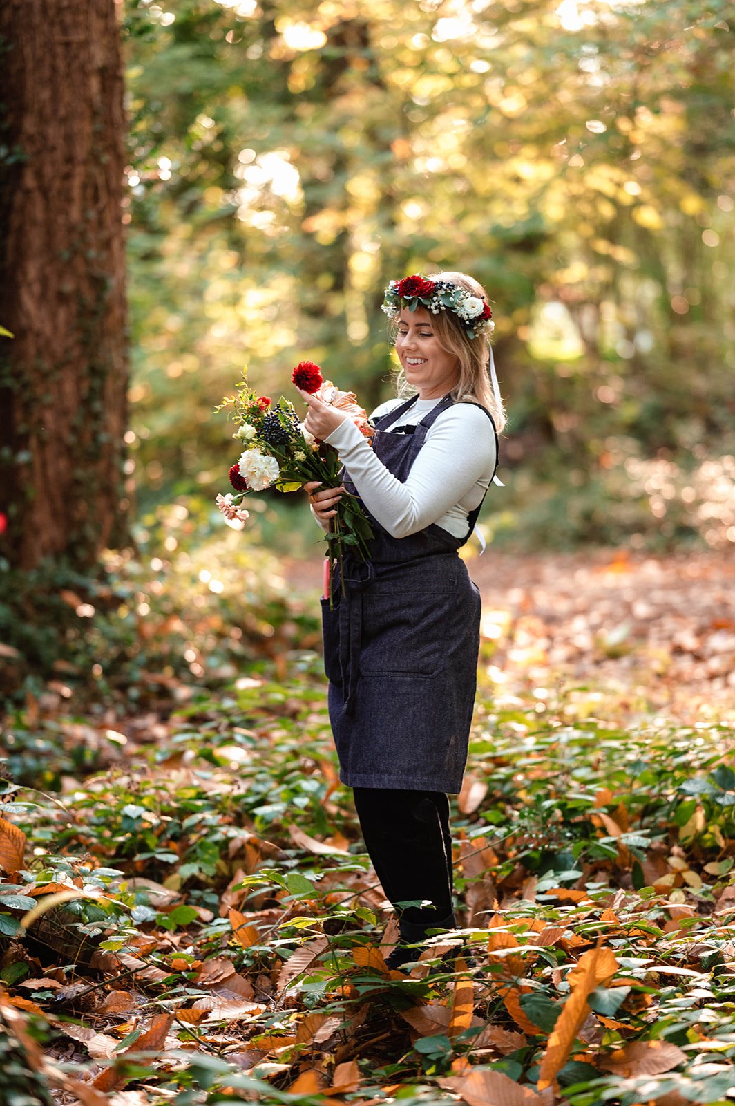 A woman in a white long-sleeve shirt and dark apron stands in a forest with autumn leaves on the ground, holding a bouquet of flowers and a cupcake, wearing a flower crown, and smiling.