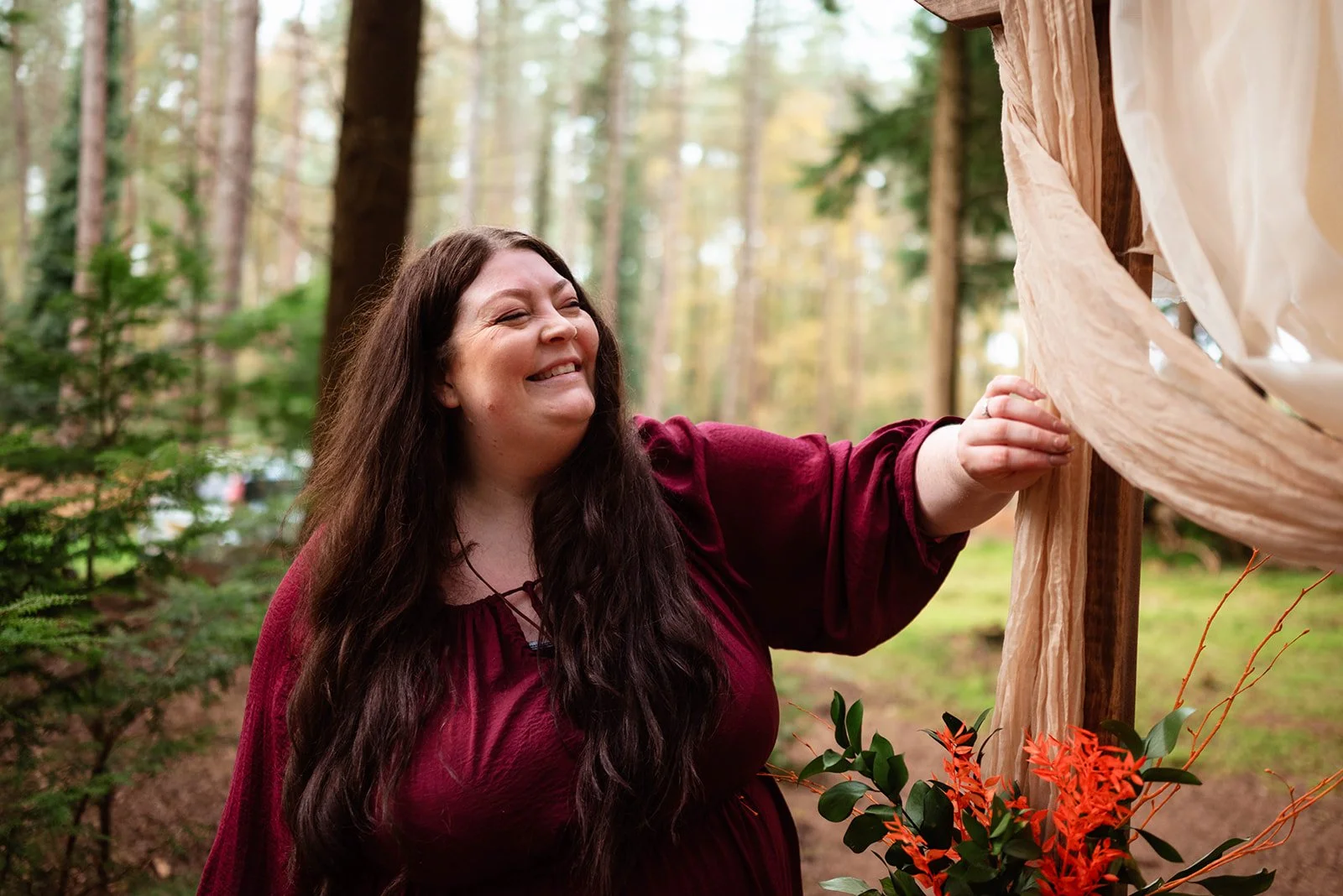 A woman with long dark hair, wearing a maroon dress, smiles happily as she touches a piece of fabric or paper outside in a wooded area during daytime.