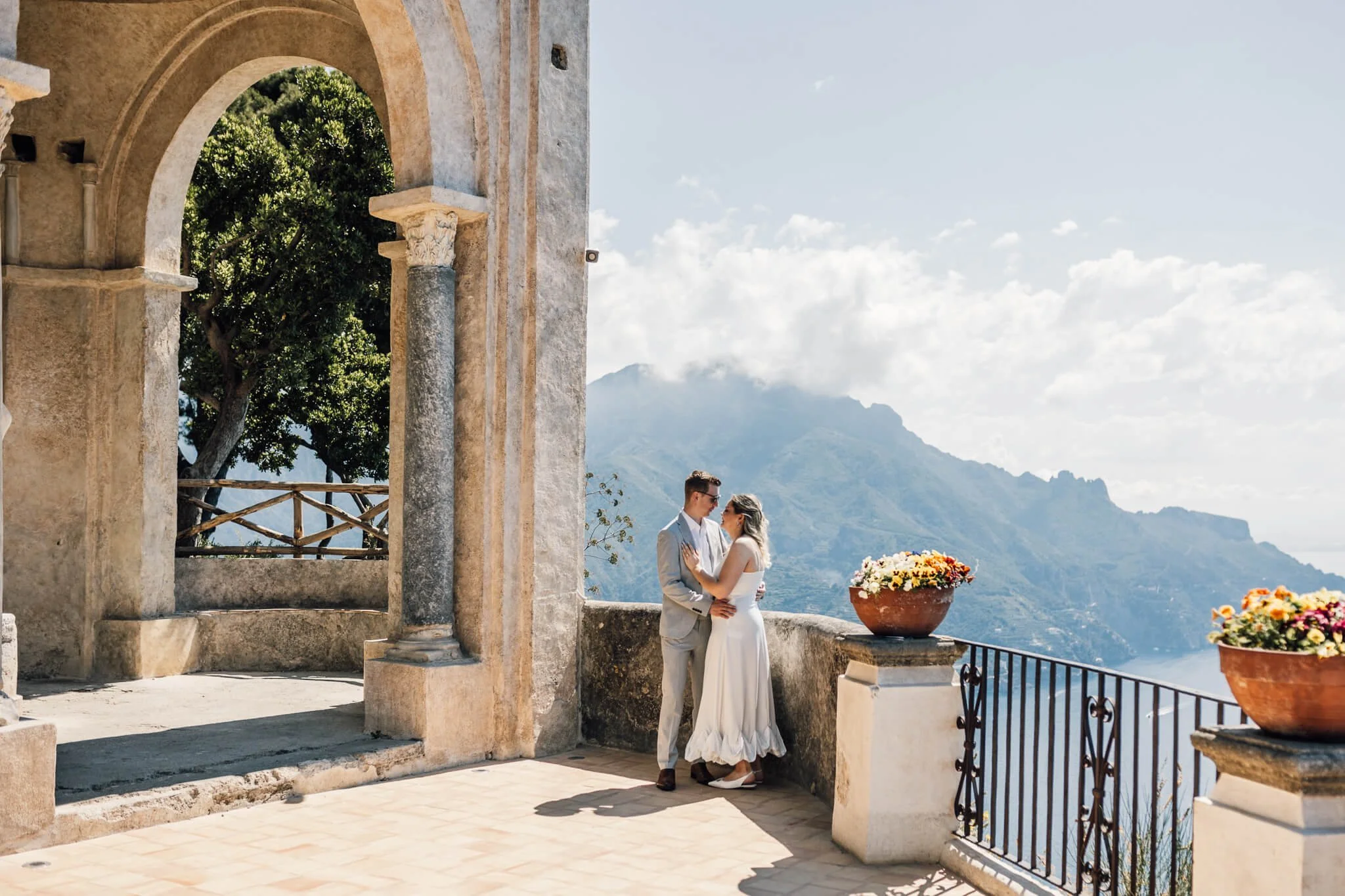 A couple dressed in wedding attire standing on a terrace with mountain views, potted flowers, and ancient stone arches.