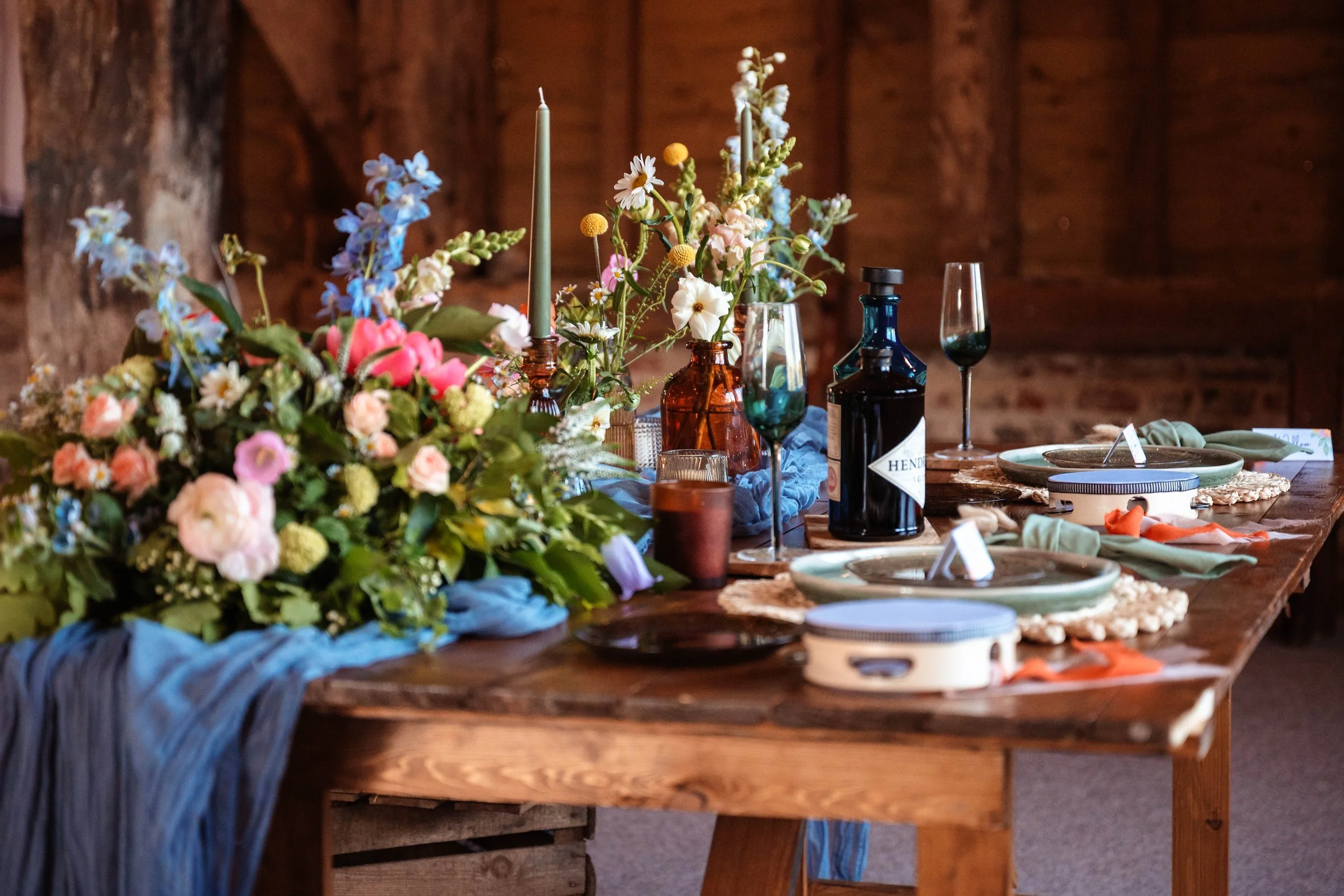 Rustic wooden table decorated with colorful flowers, candles, and glassware, set for a meal in a cozy barn-like setting.
