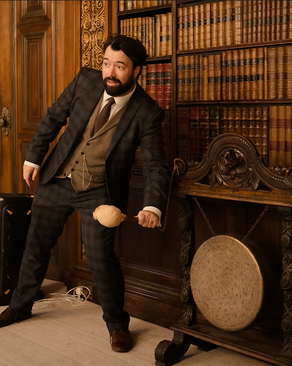 Man in a suit holding a mallet and hitting a gong in a room with wooden bookshelves filled with books.