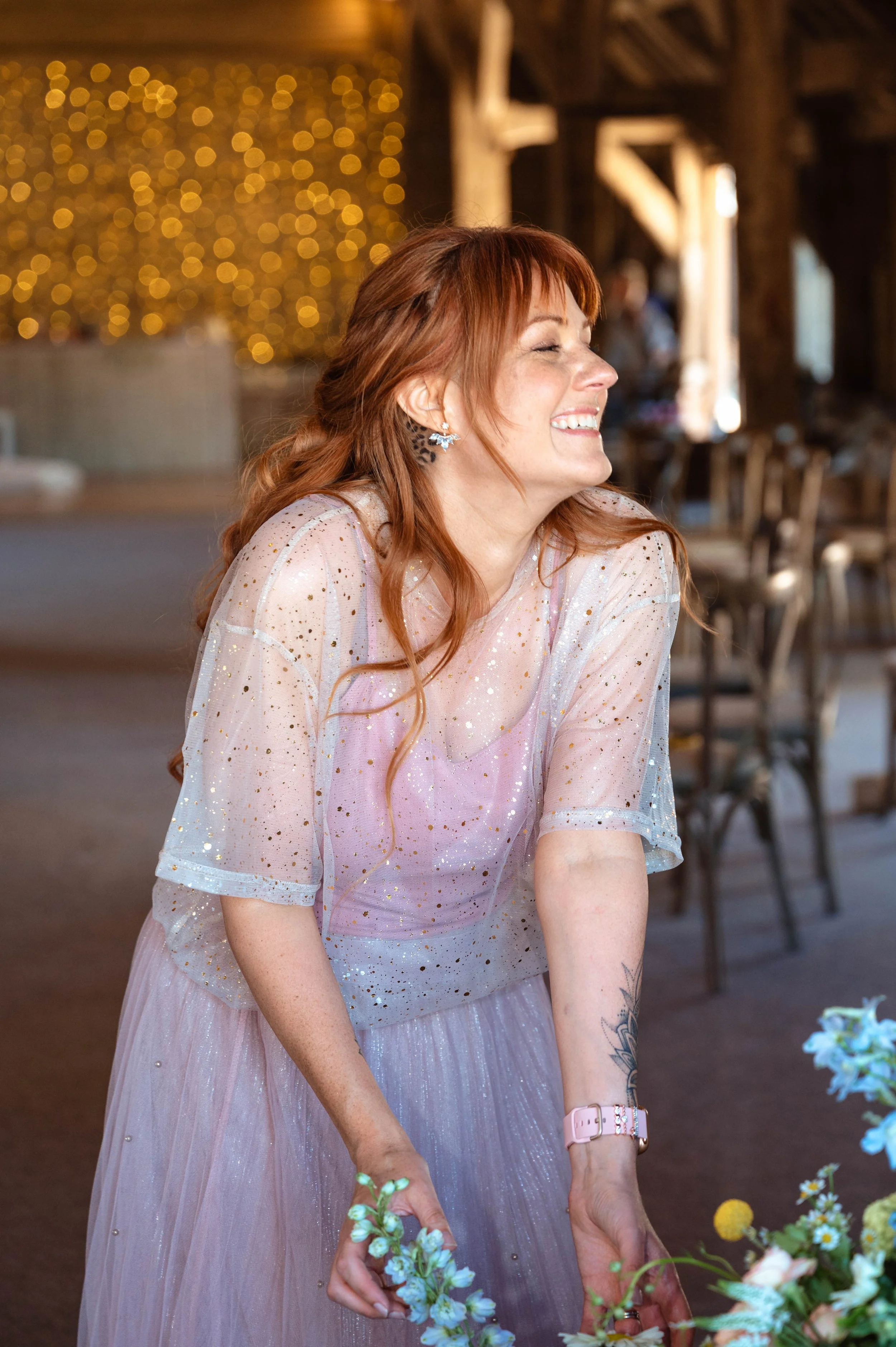A woman with long red hair and earrings, smiling happily, wearing a sheer dress with glitter, standing in a rustic venue decorated with fairy lights.