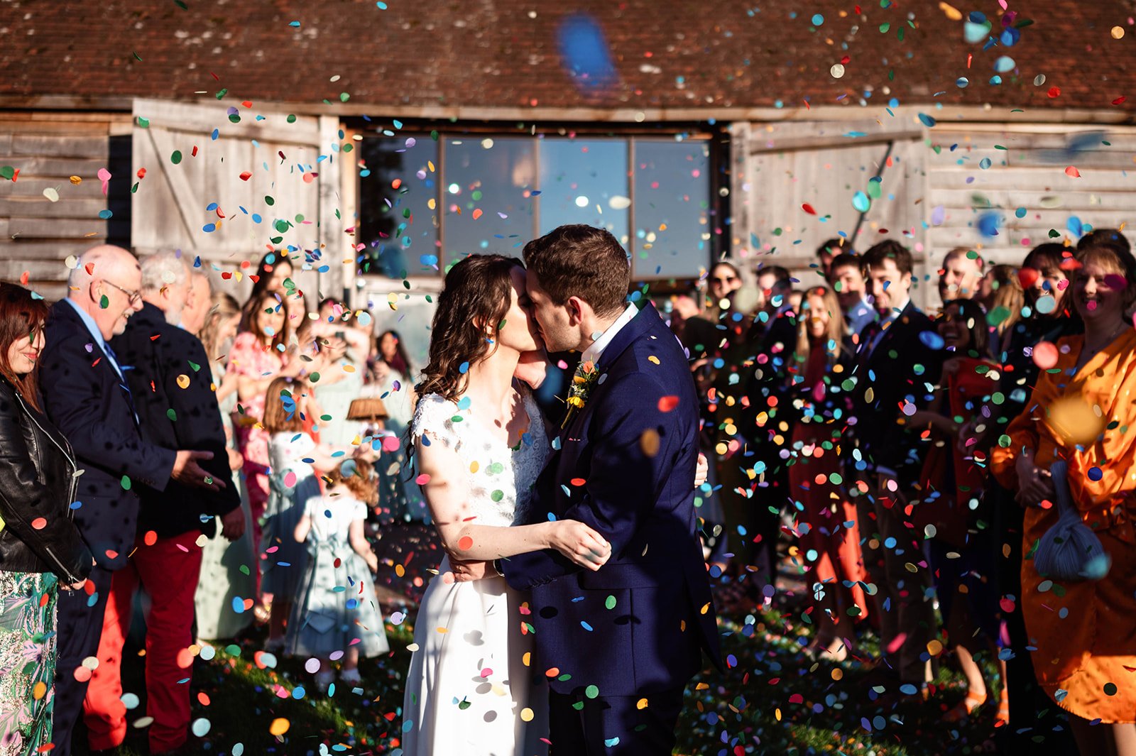 A newlywed couple kissing while surrounded by a crowd at a wedding celebration, with colorful confetti falling from above outside a rustic wooden barn.
