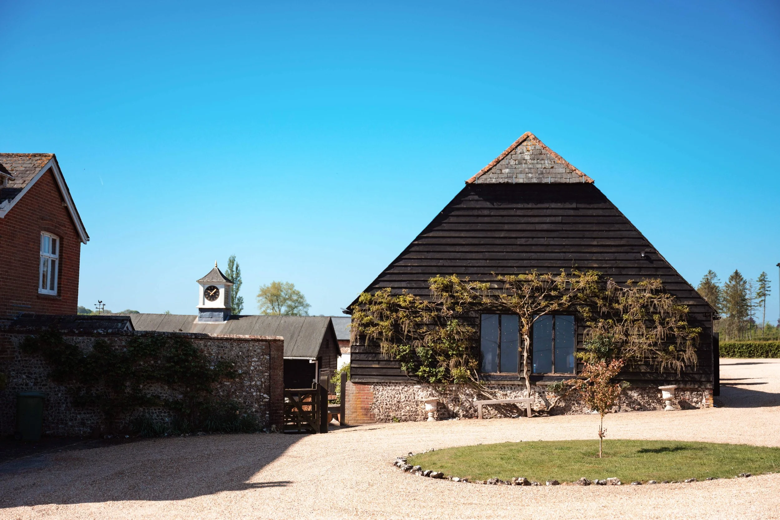A rustic rural scene featuring a black wooden building with a gabled roof, a small grassy area with a young tree, a gravel pathway, and part of another brick building with a white steeple in the background under a clear blue sky.