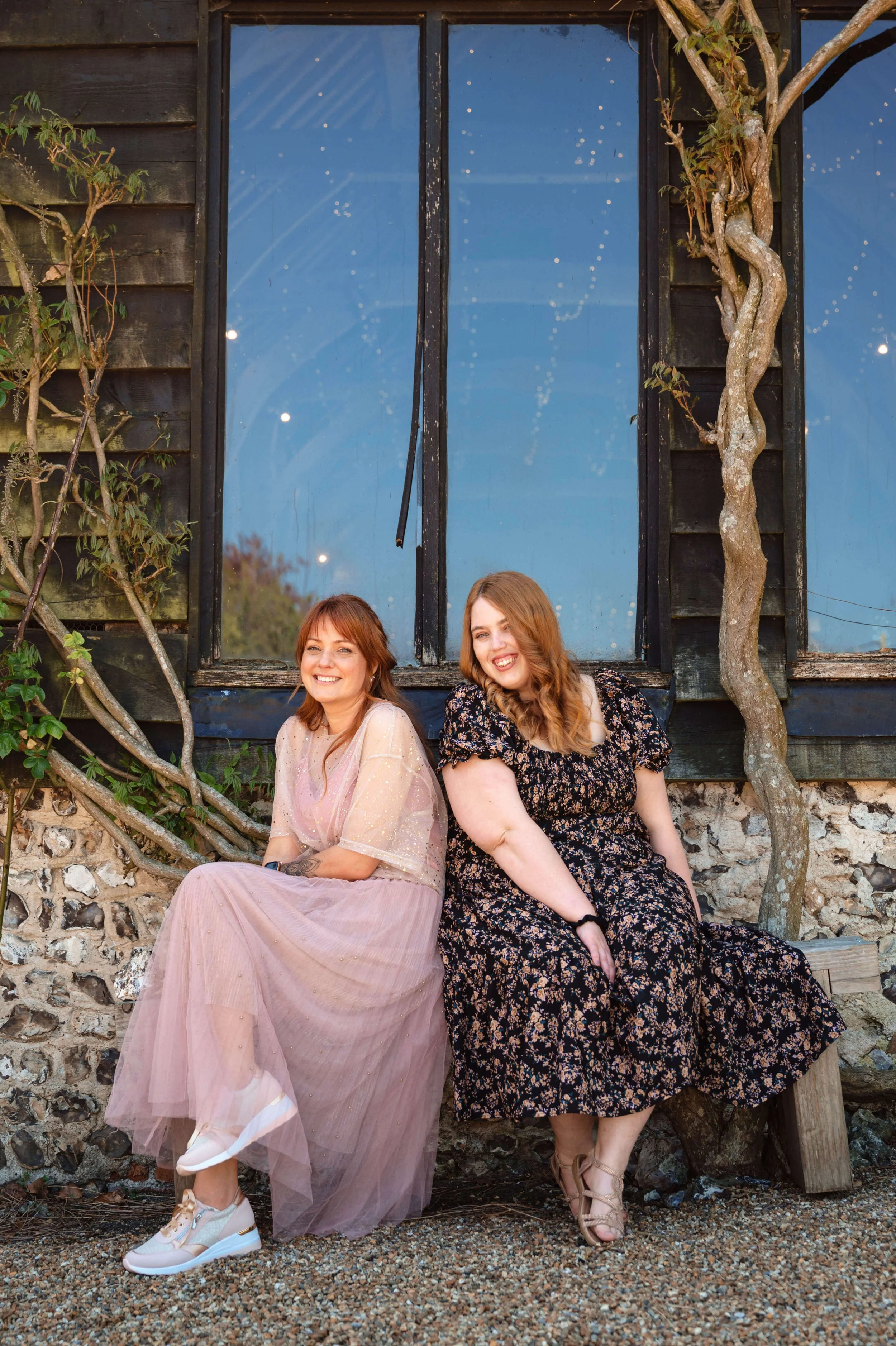 Two women sitting on a wooden bench outside, smiling at the camera, with a building and a large window behind them and some climbing plants.