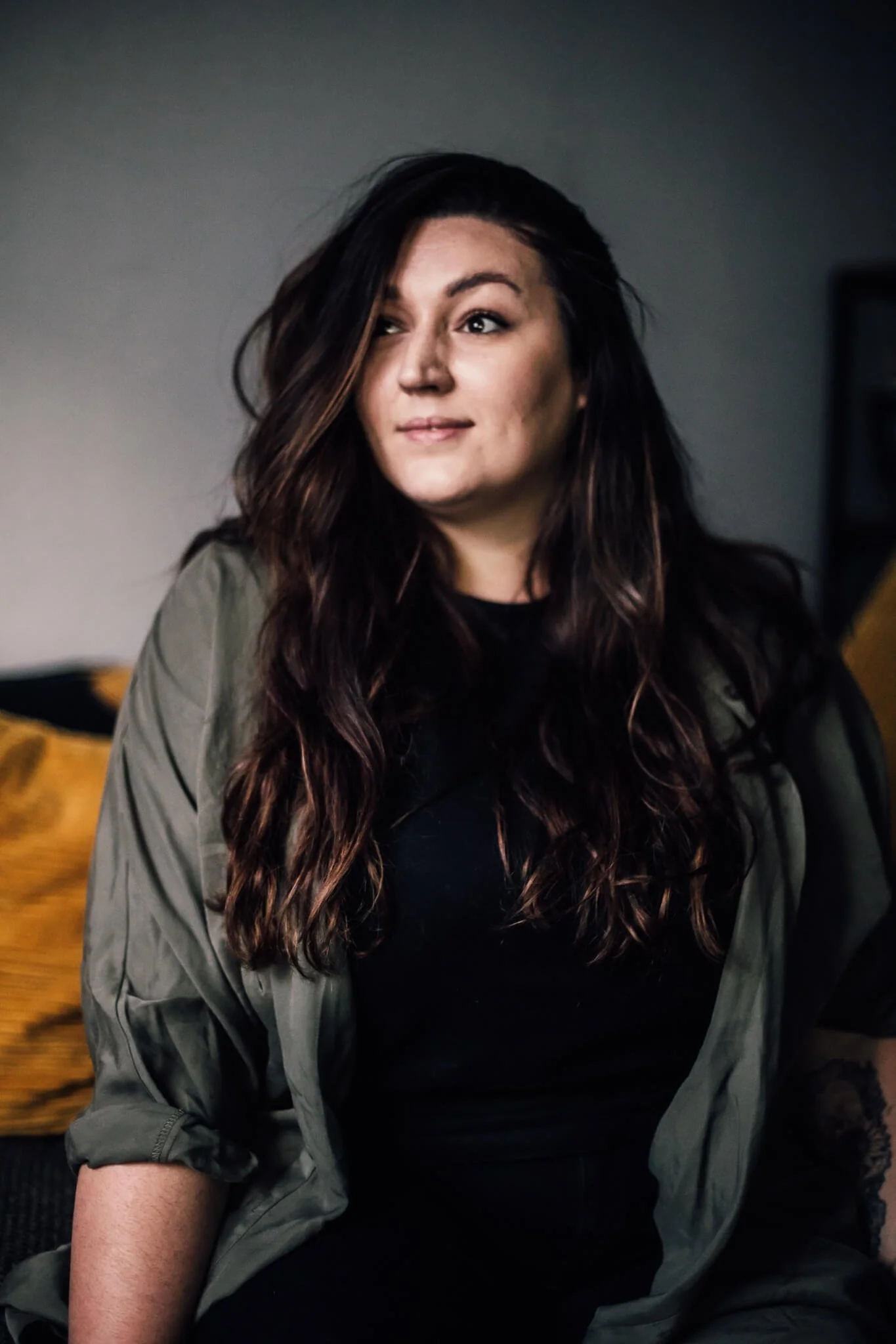 A young woman with long wavy brown hair, wearing a dark shirt and an olive green jacket, sitting indoors with a neutral expression, and a yellow pillow in the background.