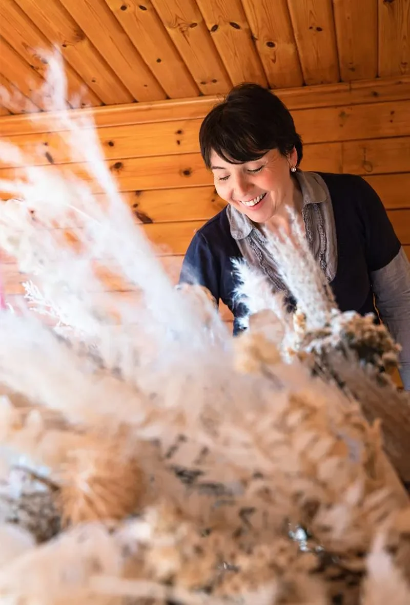 A woman with short dark hair smiling indoors with wooden walls behind her, looking down at a fluffy animal, possibly a dog with a black nose, on a log or similar surface.