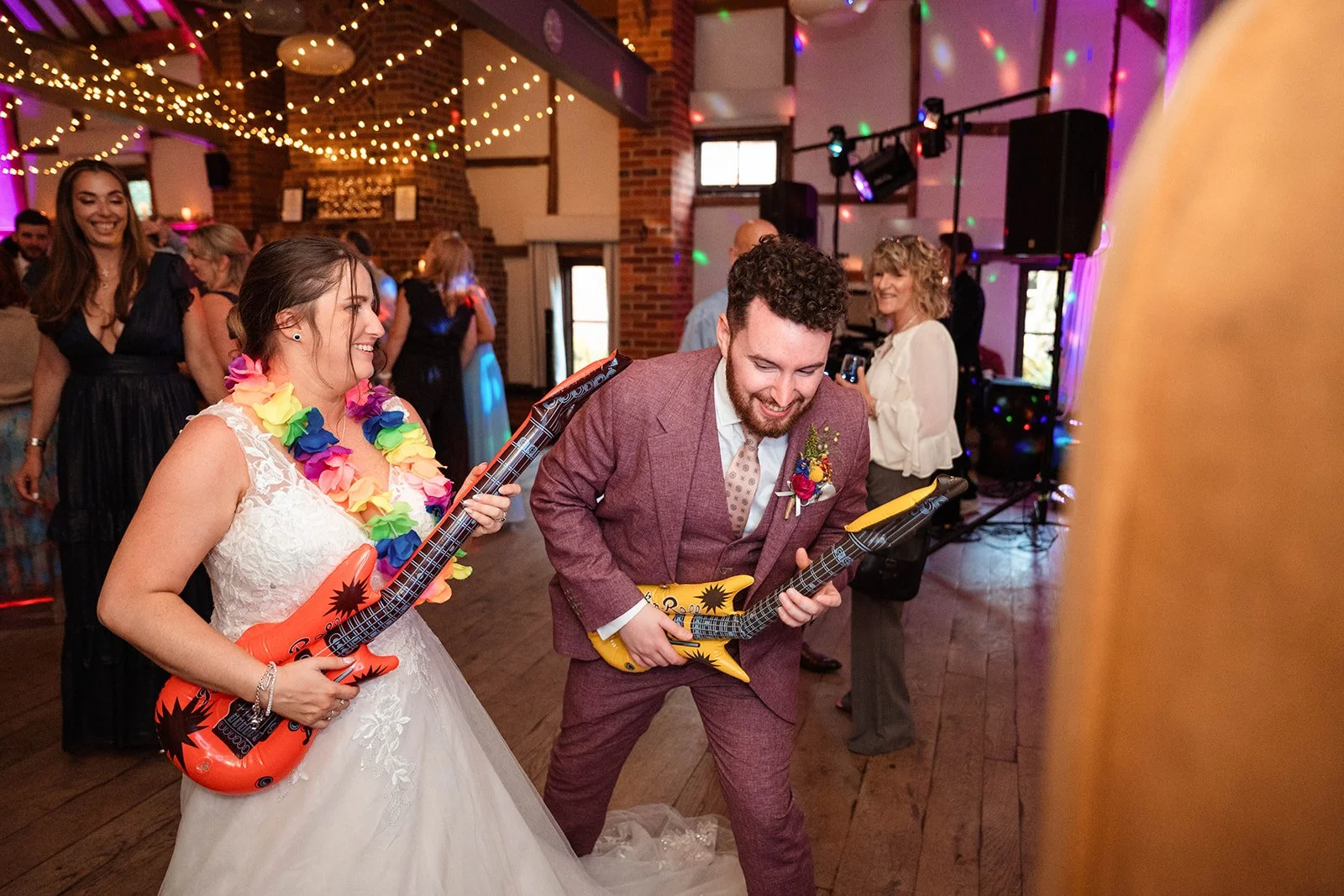 Bride and groom playing inflatable guitars at wedding reception with guests dancing and decorated venue with string lights.