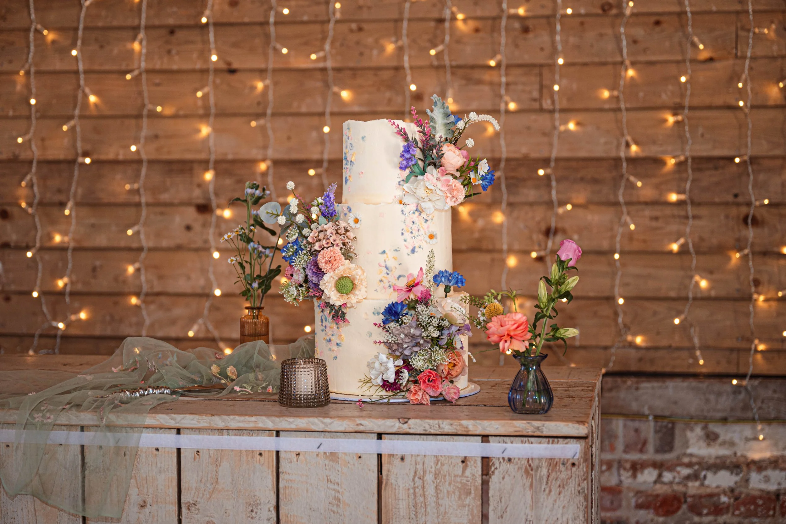 A tall, white wedding cake decorated with colorful flowers, placed on a rustic wooden table, with small vases of flowers and decorative candles around it. The background features wooden planks with string lights.