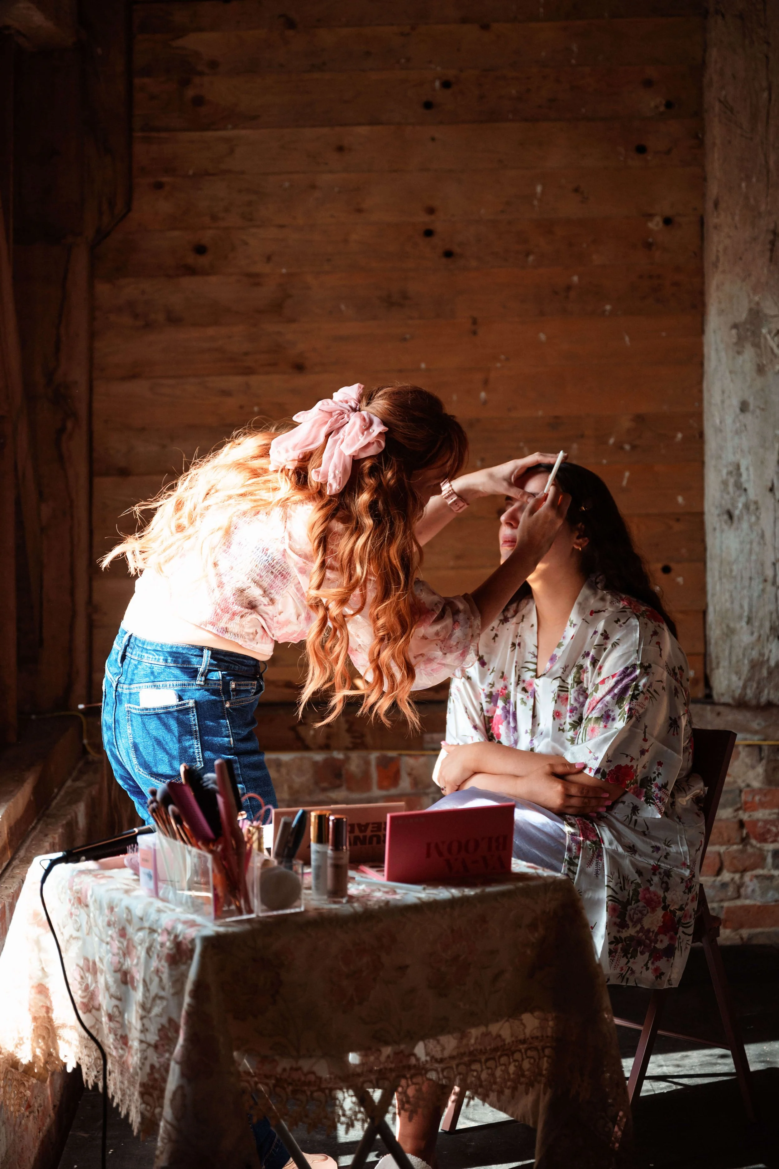 A makeup artist applying makeup to a woman seated in front of a rustic wooden wall, with a table of beauty supplies in front of them.