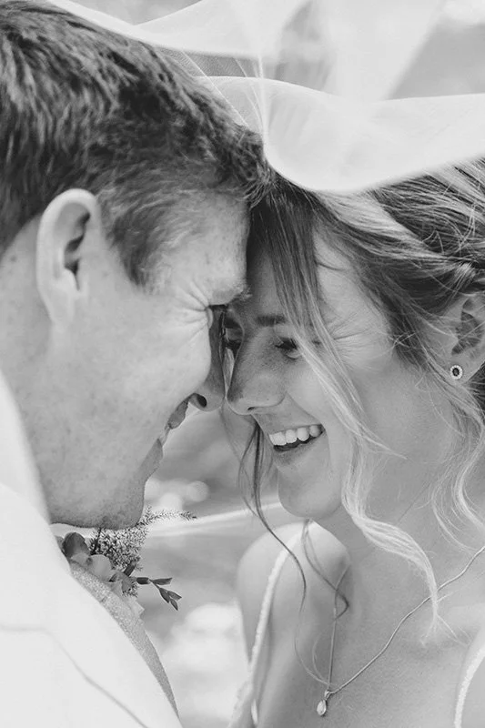 A couple on their wedding day, smiling and touching foreheads under a veil, in black and white. border
