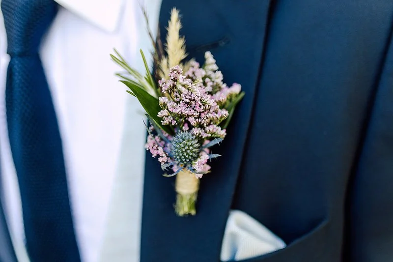 Close-up of a boutonniere with pink and purple flowers and greenery pinned to a dark suit jacket. border