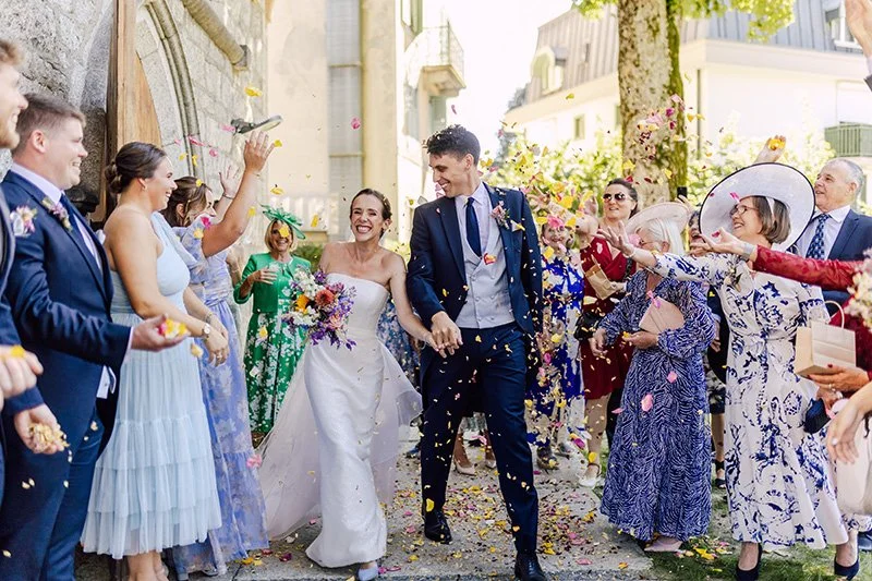 Bride and groom walking through a shower of colorful flower petals and confetti, surrounded by wedding guests in Annecy. border