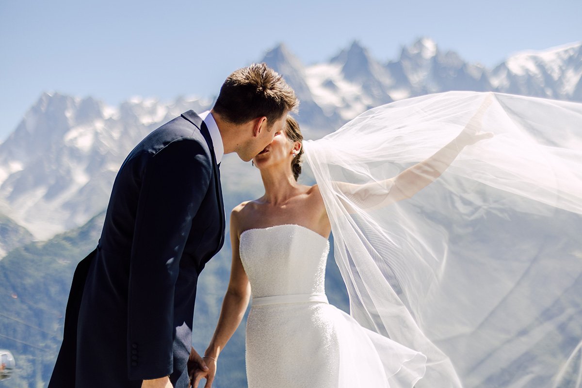Bride and groom kissing outdoors in a mountainous area with snow-capped Chamonix peaks in the background. The bride is wearing a strapless white wedding gown and veil, and the groom is dressed in a dark suit. border