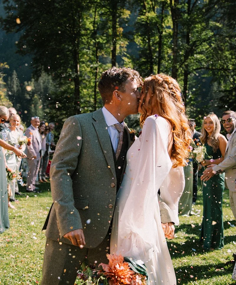 Romantic kiss between bridge and groom at their Domaine du Baron Lake Montriond wedding ceremony, with wedding guests celebrating in the background and confetti border