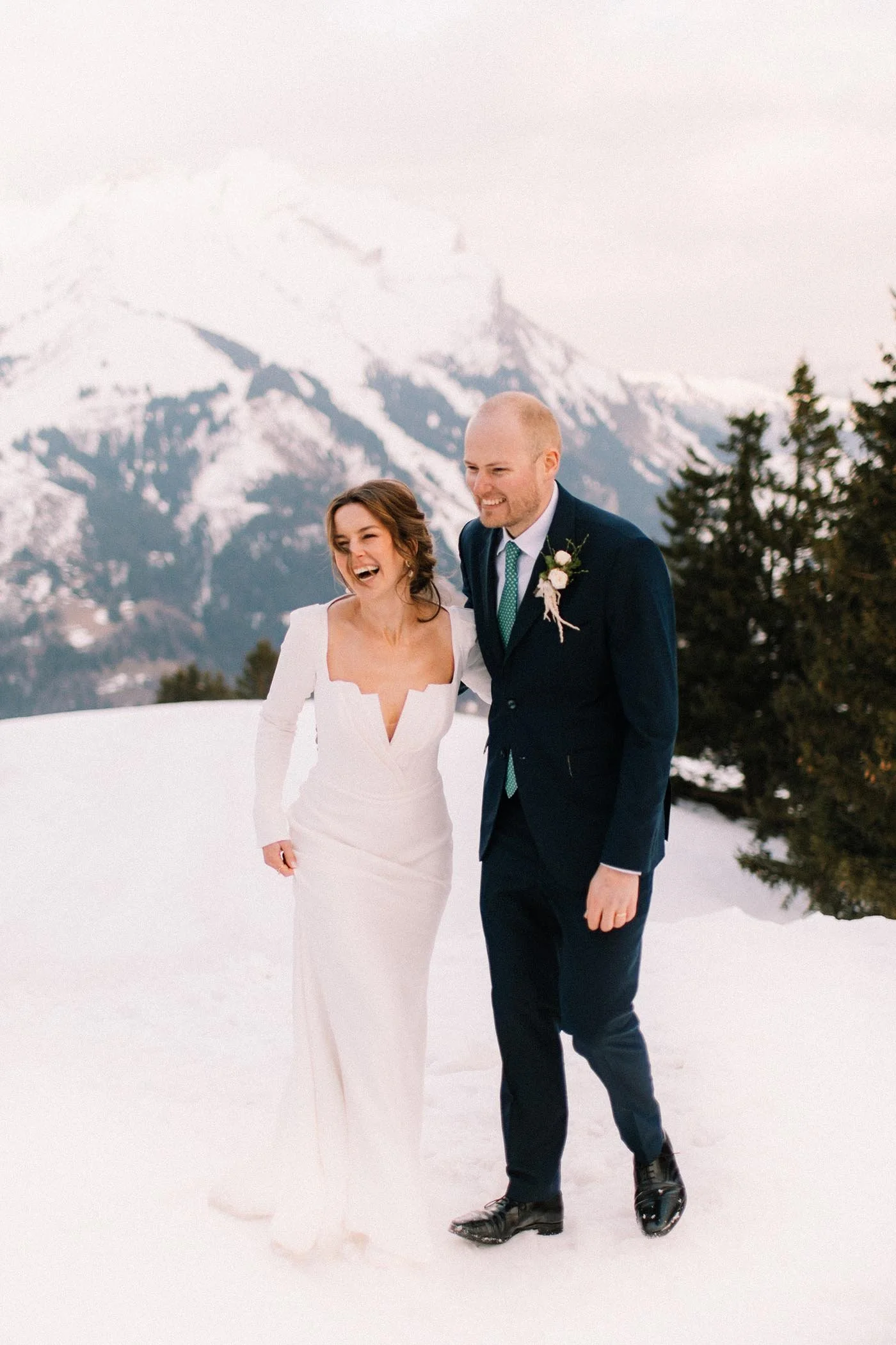 A bride and groom walk together on snow-covered ground with snow-capped mountains in the background. The bride is in a white wedding dress, and the groom is in a navy suit with a boutonniere, wearing ice skates border