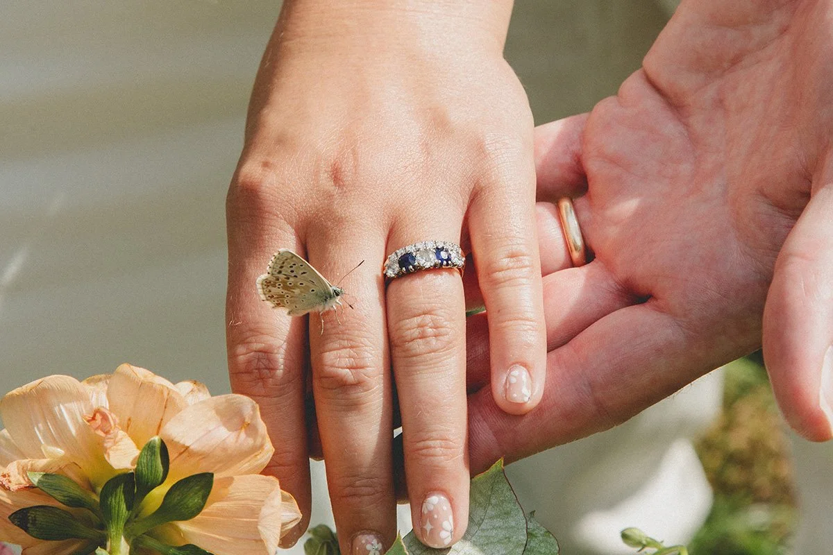 Close-up of two hands, one with a butterfly on a finger, and the other with rings, surrounded by flowers and greenery. border