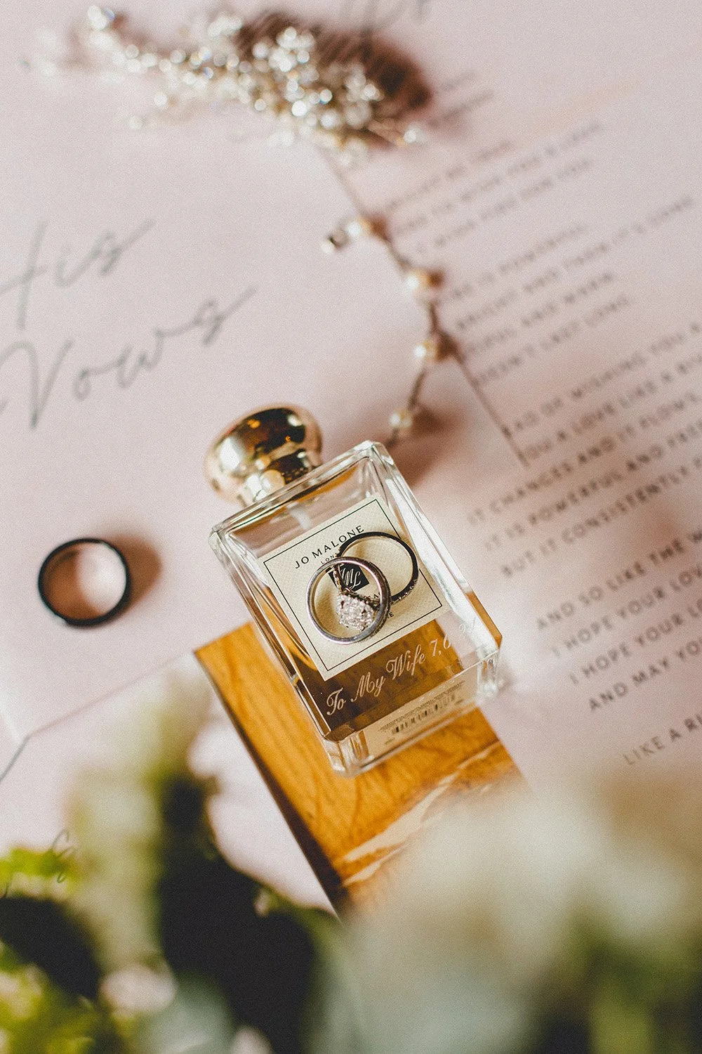 A perfume bottle with a diamond engagement ring on top surrounded by wedding rings, set on a wooden block with a pink note and a letter underneath, with blurred flowers in the foreground. border