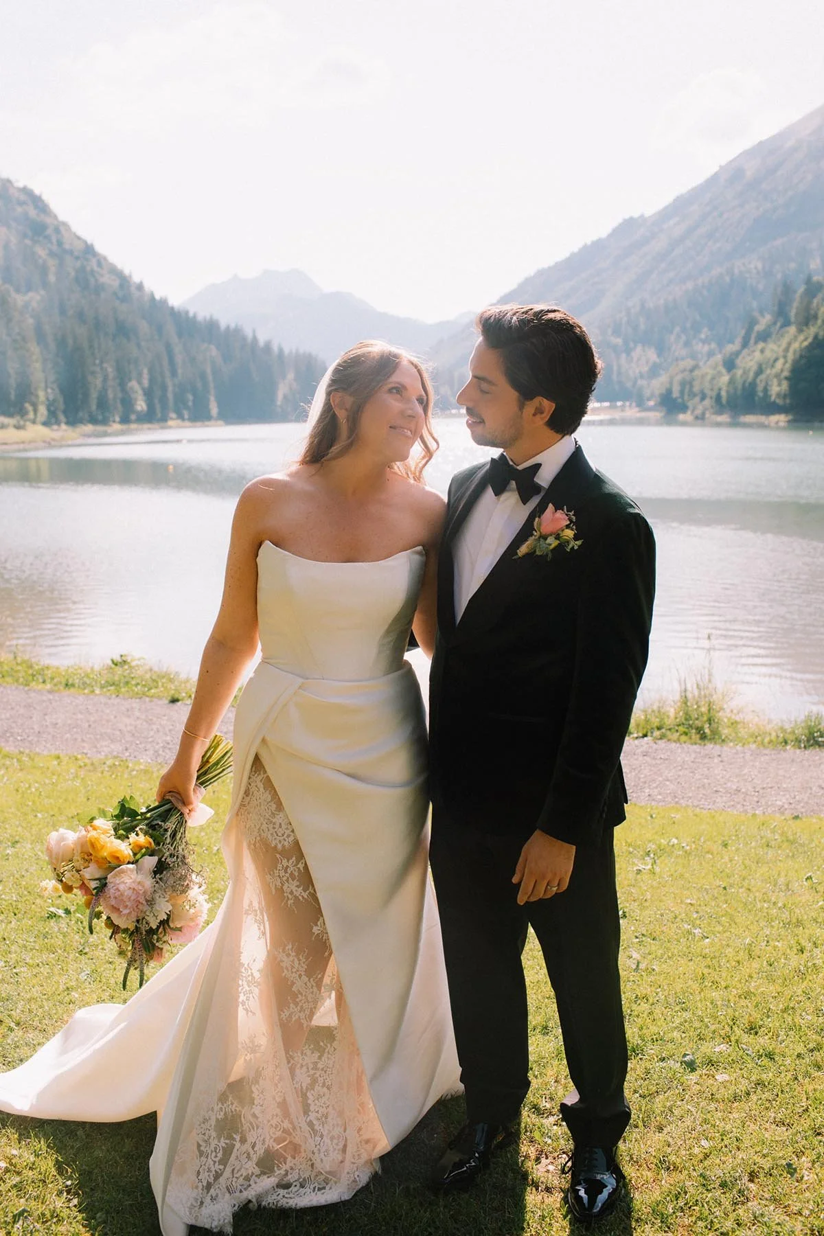A bride in a strapless white wedding gown holding a bouquet and a groom in a black tuxedo with a pink boutonniere standing by Lake Montriond border