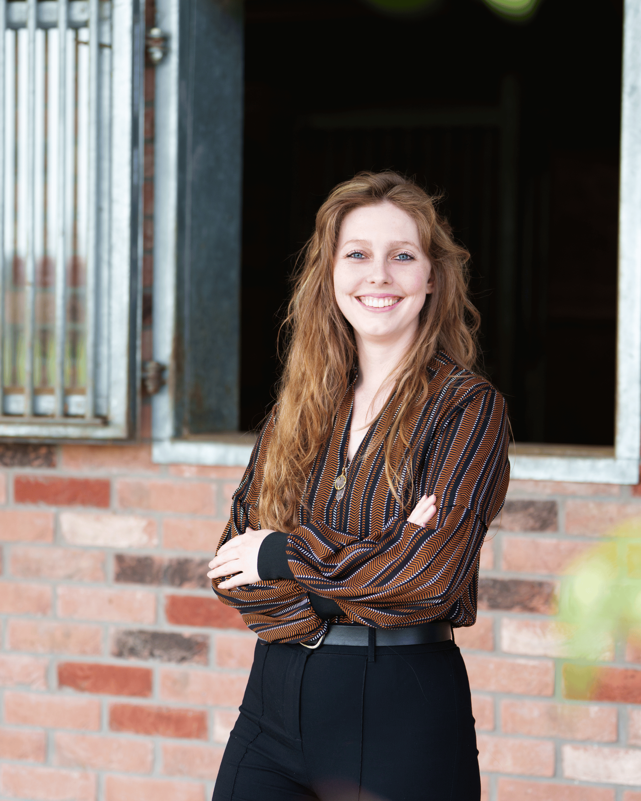 Woman with long hair smiling in front of a brick wall, wearing a striped blouse and black pants.