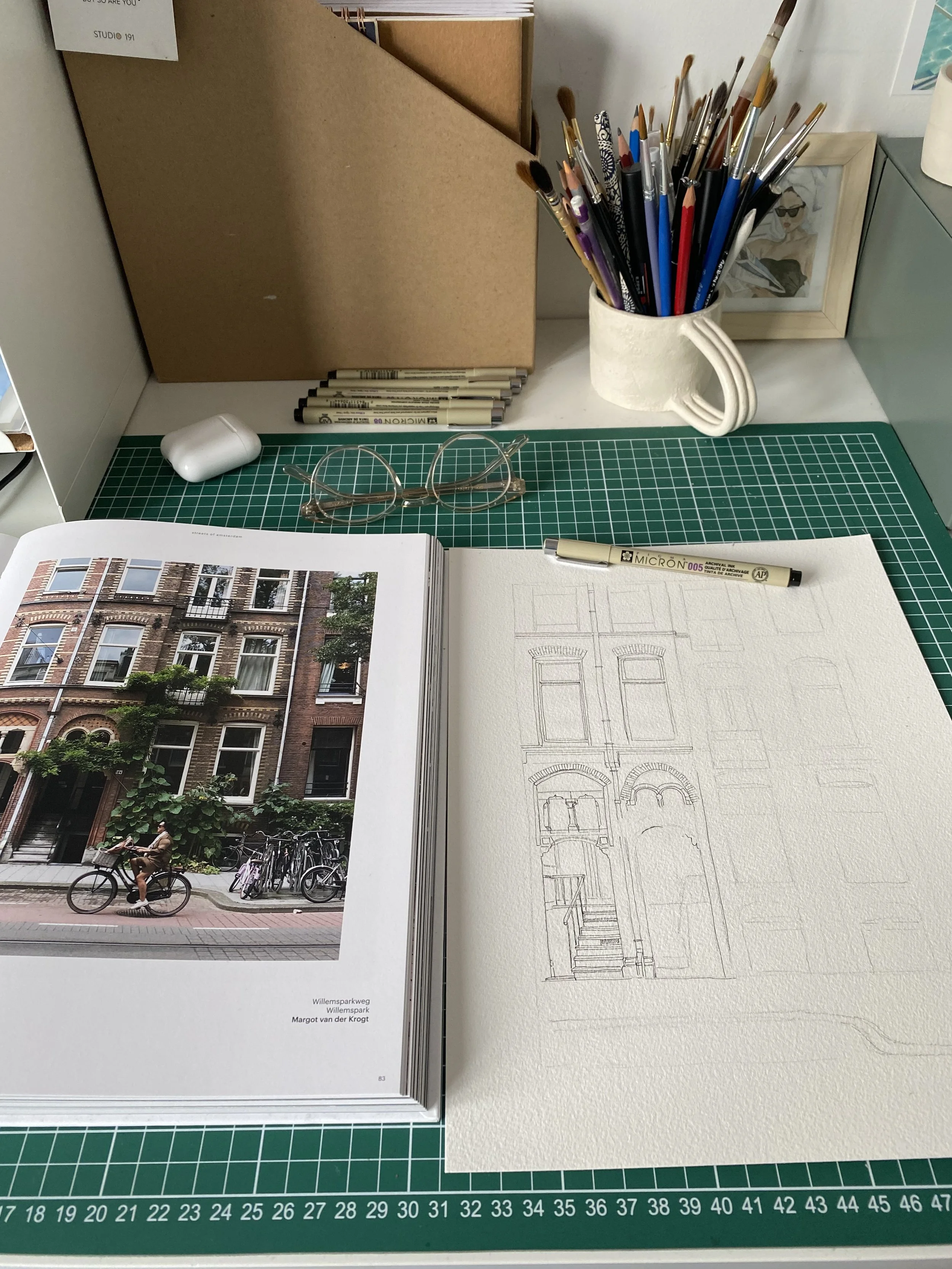A workspace with a photo book open showing a building facade and a bicycle, a sketch of building windows and doorways on textured paper, a green cutting mat, a pair of glasses, a white AirPods case, a container of paintbrushes, a small framed photo, and a marker on a desk.