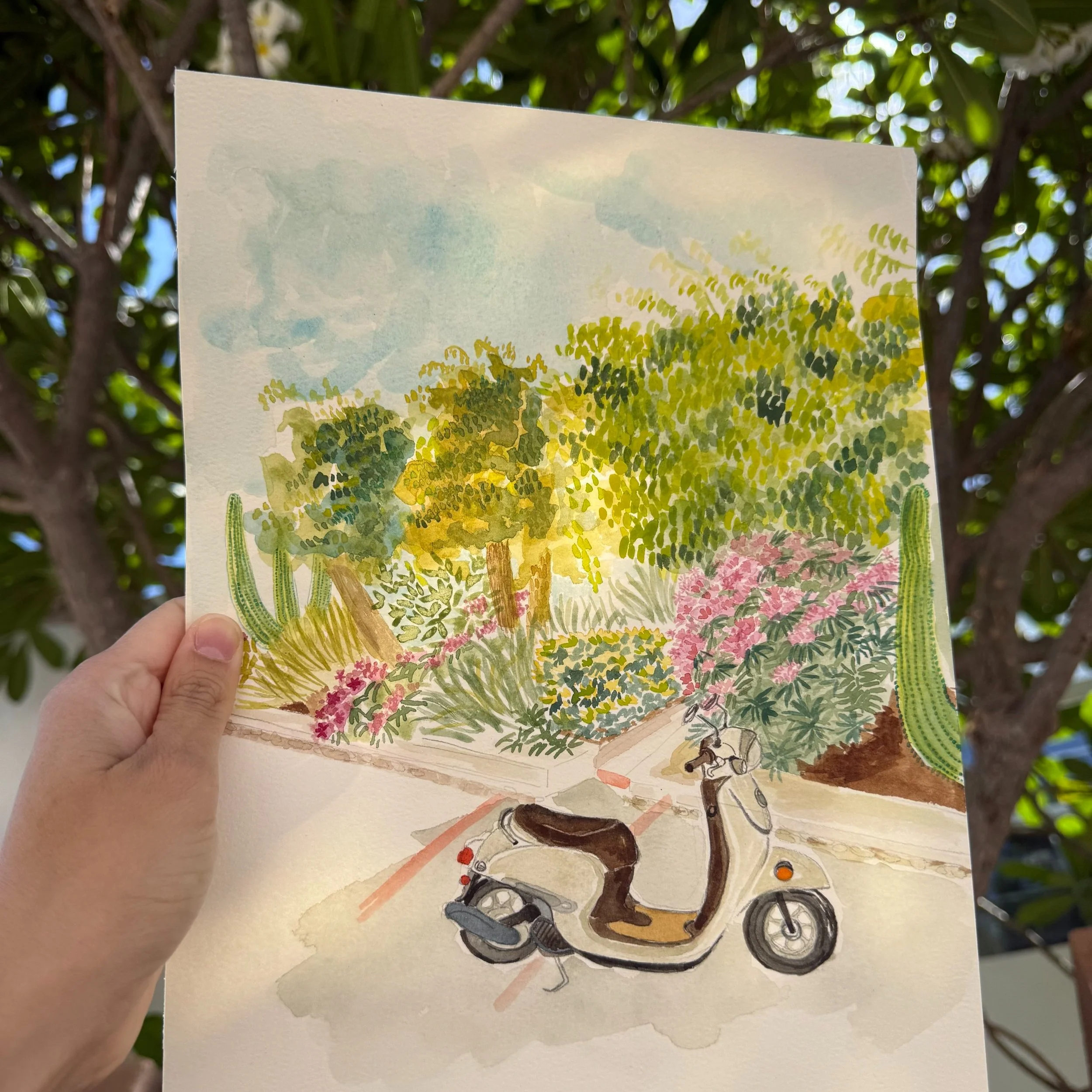 A hand holding a watercolor painting of a scooter parked on a sidewalk, surrounded by lush green trees, flowering bushes, and cacti, with a blue sky in the background.