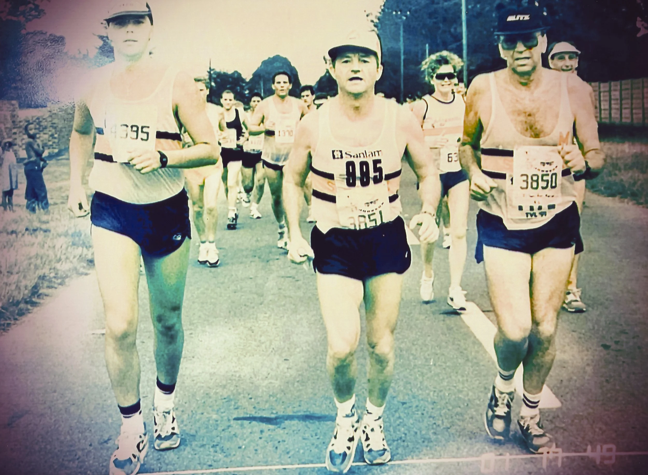 Fred and a group of runners participating in a marathon, wearing athletic gear and bib numbers, running on a paved road with trees and a fence in the background.