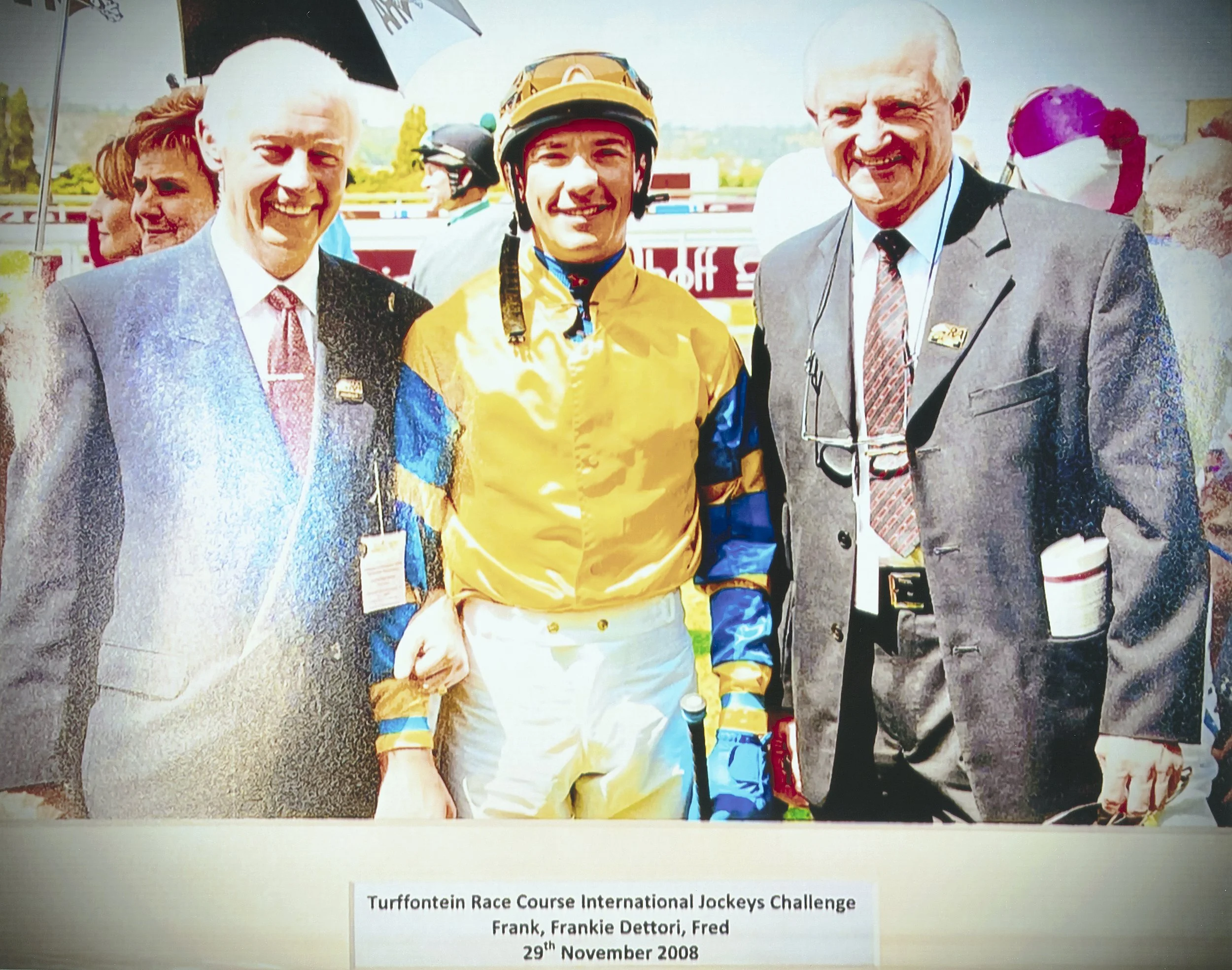 Frank, Frankie Dettori, and Fred standing outdoors at a horse racing event, with a racing jockey in racing gear and two men in suits. The background shows other people and racing banners.