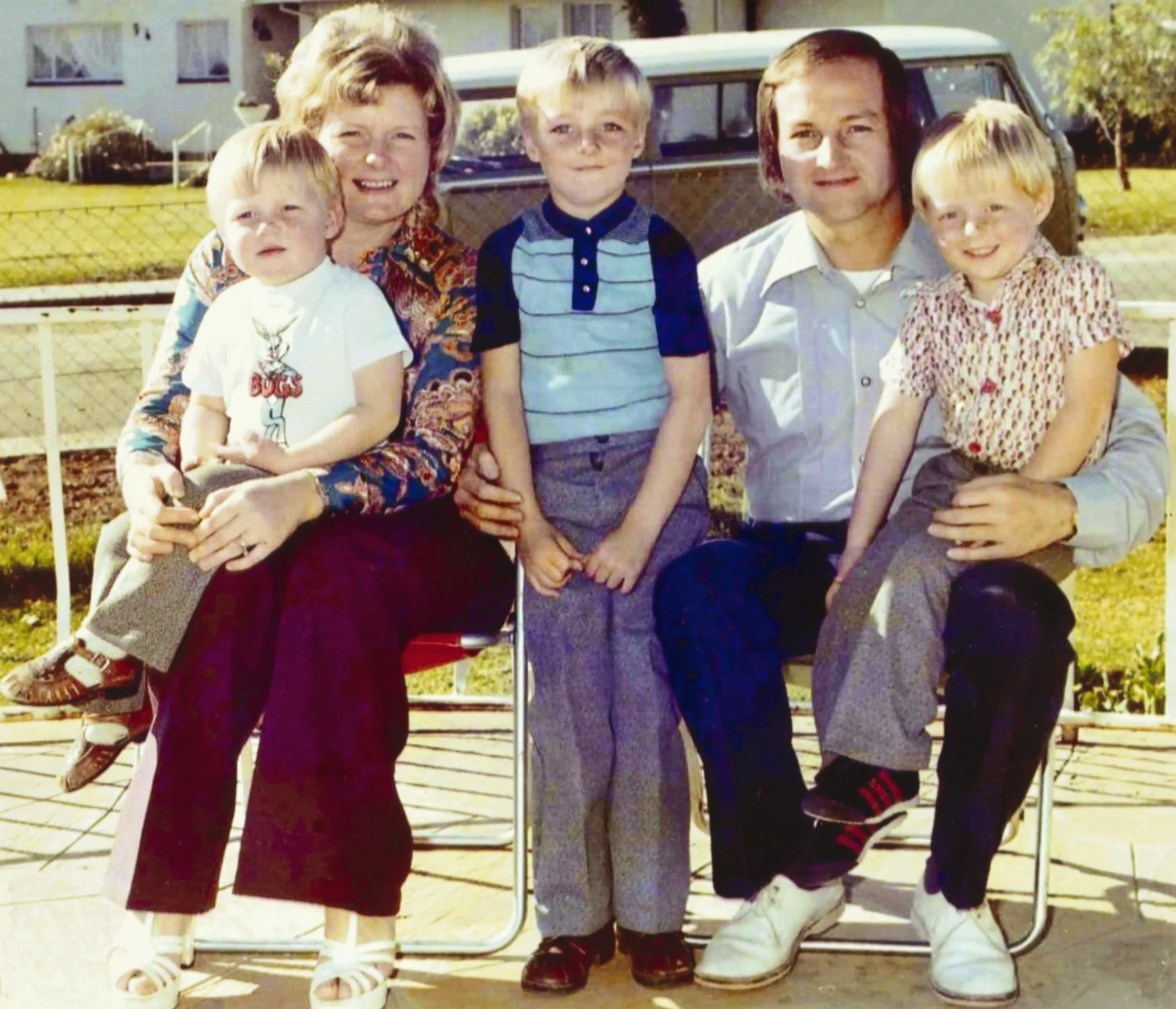 The Thompson family posing outdoors on a sunny day, sitting on a wooden deck with a metal fence and a vintage van in the background.