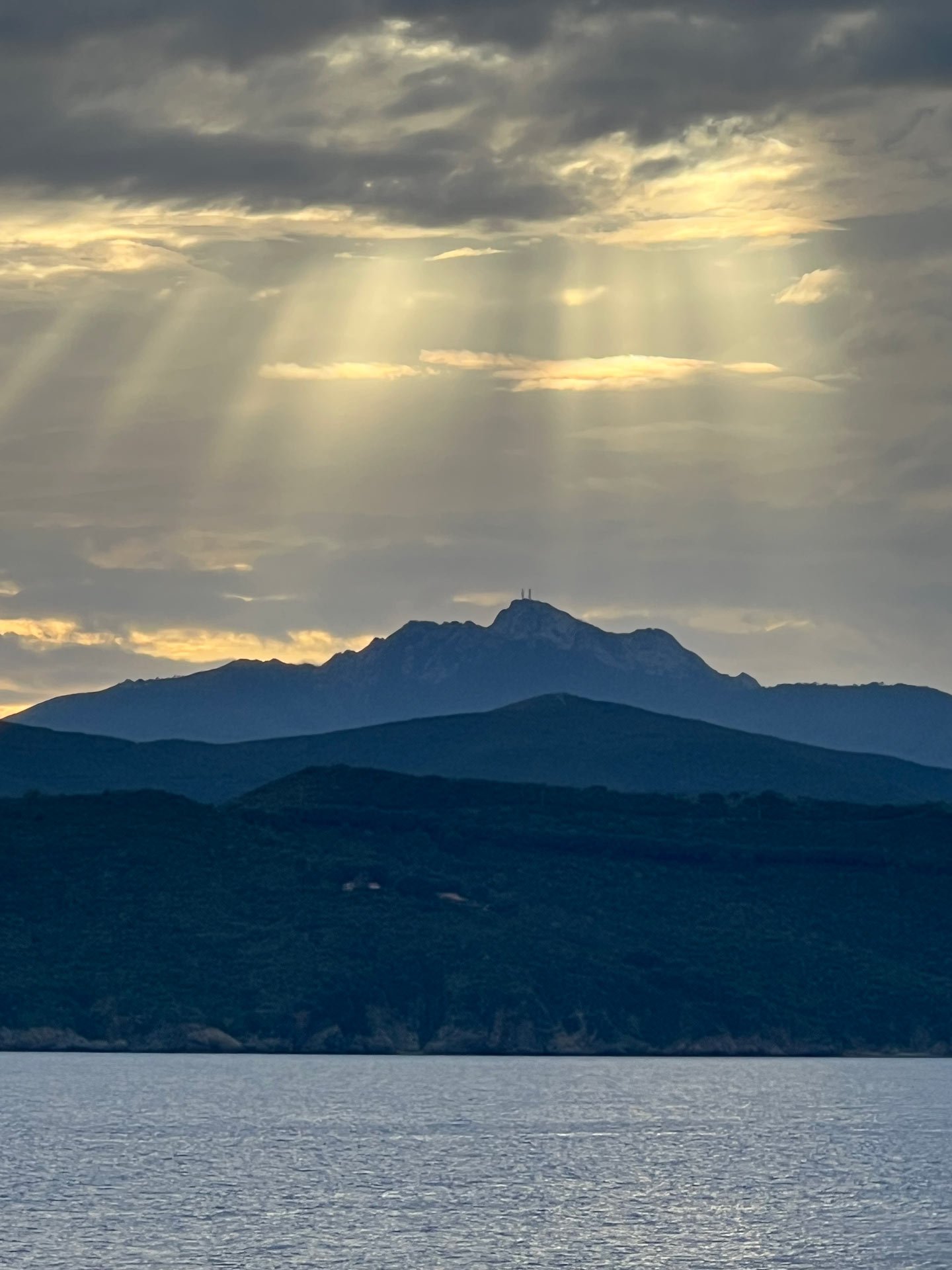 Berglandschaft mit einem See im Vordergrund und bewölktem Himmel mit Sonnenstrahlen.