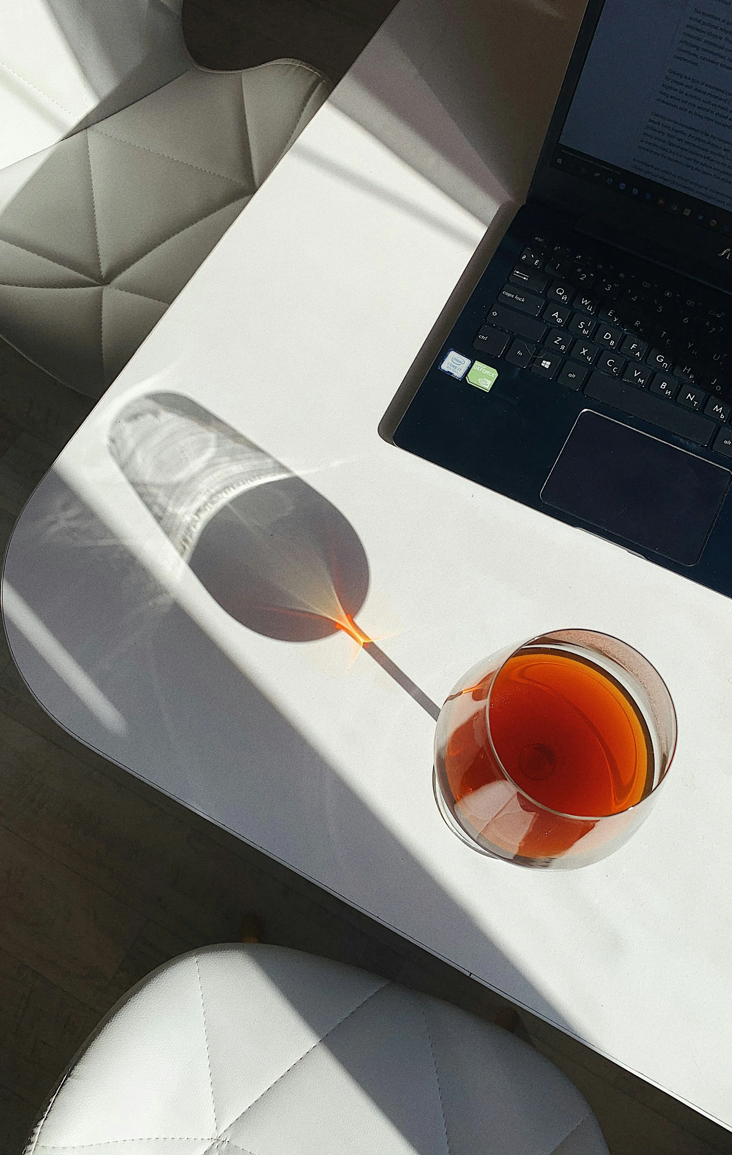 Ein weißer Tisch mit einem Laptop, einem Glas Tee und einem Schatten, der den Verlauf einer Papiertüte auf der Tischfläche zeigt, in Sonnenlicht.