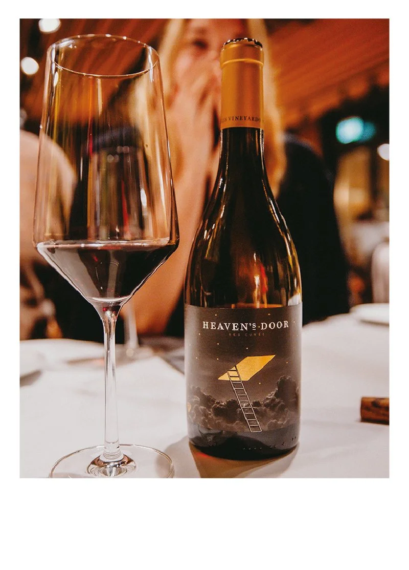 Weinglas mit rotem Wein neben einer Flasche mit einem Himmel-Design und der Aufschrift 'HEAVEN'S DOOR' auf einem Tisch in einem Restaurant.