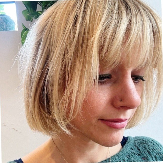 A man with curly dark hair, glasses, and a beard, wearing a dark green sweater, sitting indoors in front of a white wall with a plant visible in the background.