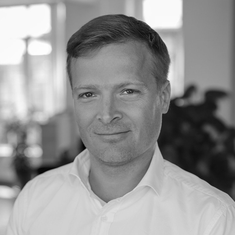 A man with dark curly hair, glasses, a nose ring, and a beard, wearing a dark green sweater, sitting indoors with a neutral background and a blurred plant in the background.