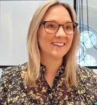 Smiling woman with shoulder-length black hair, wearing a blue top, standing in a greenhouse with green plants.