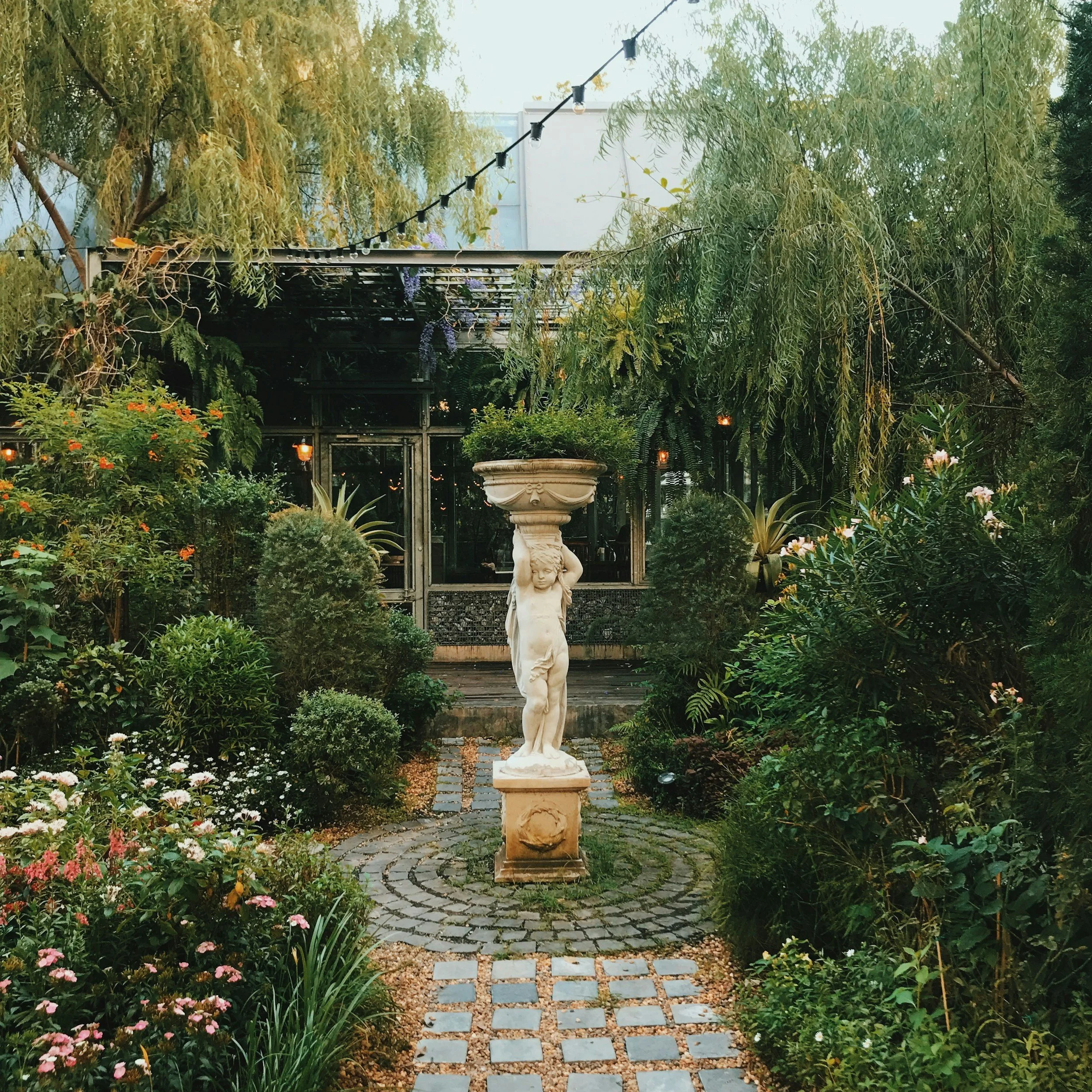 A garden with a statue of a cherub holding up a large stone bowl, surrounded by lush green plants and flowers.