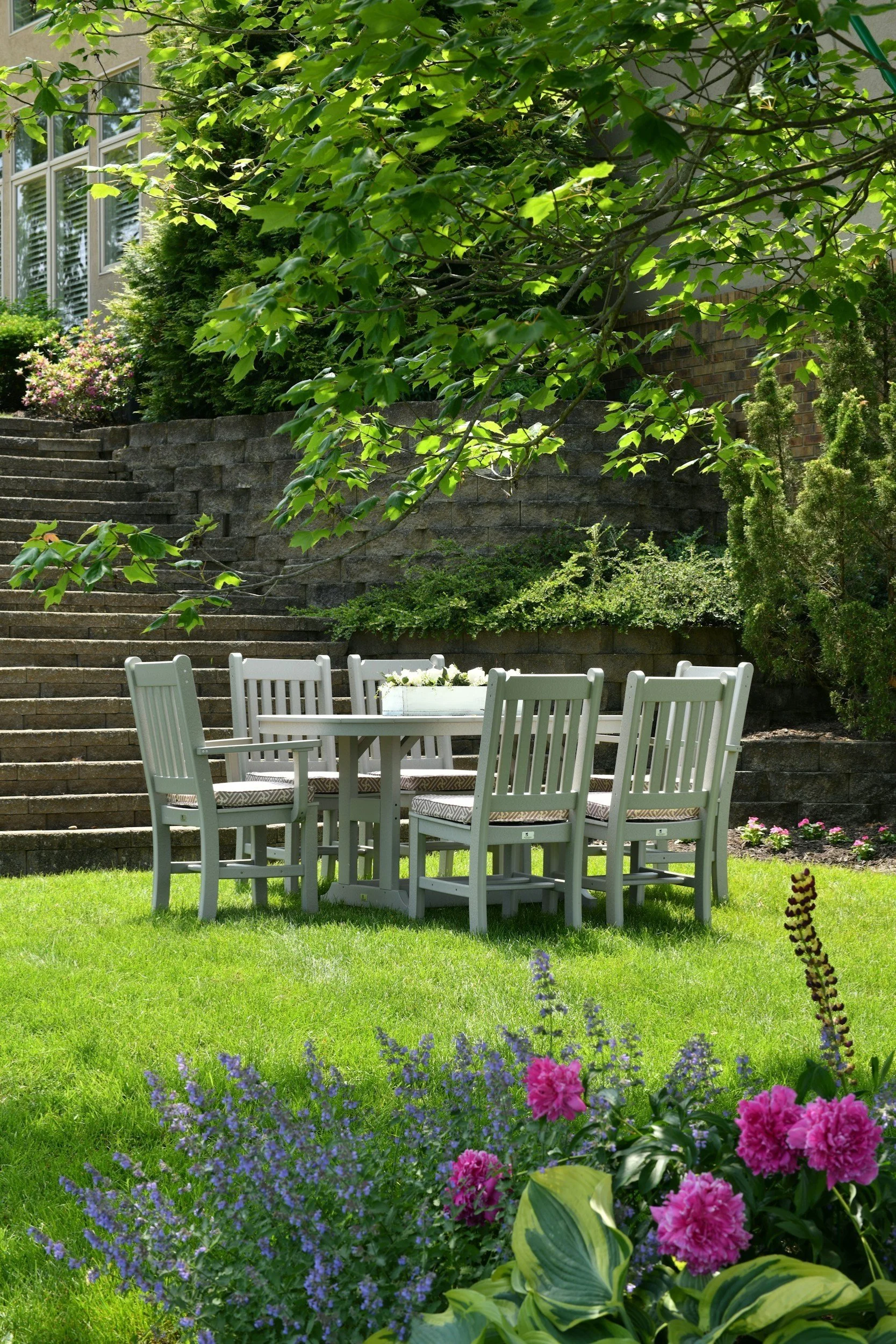 A backyard scene with a white patio table and chairs on a green lawn, surrounded by colorful flowers and lush greenery, with stone stairs and a brick wall in the background.