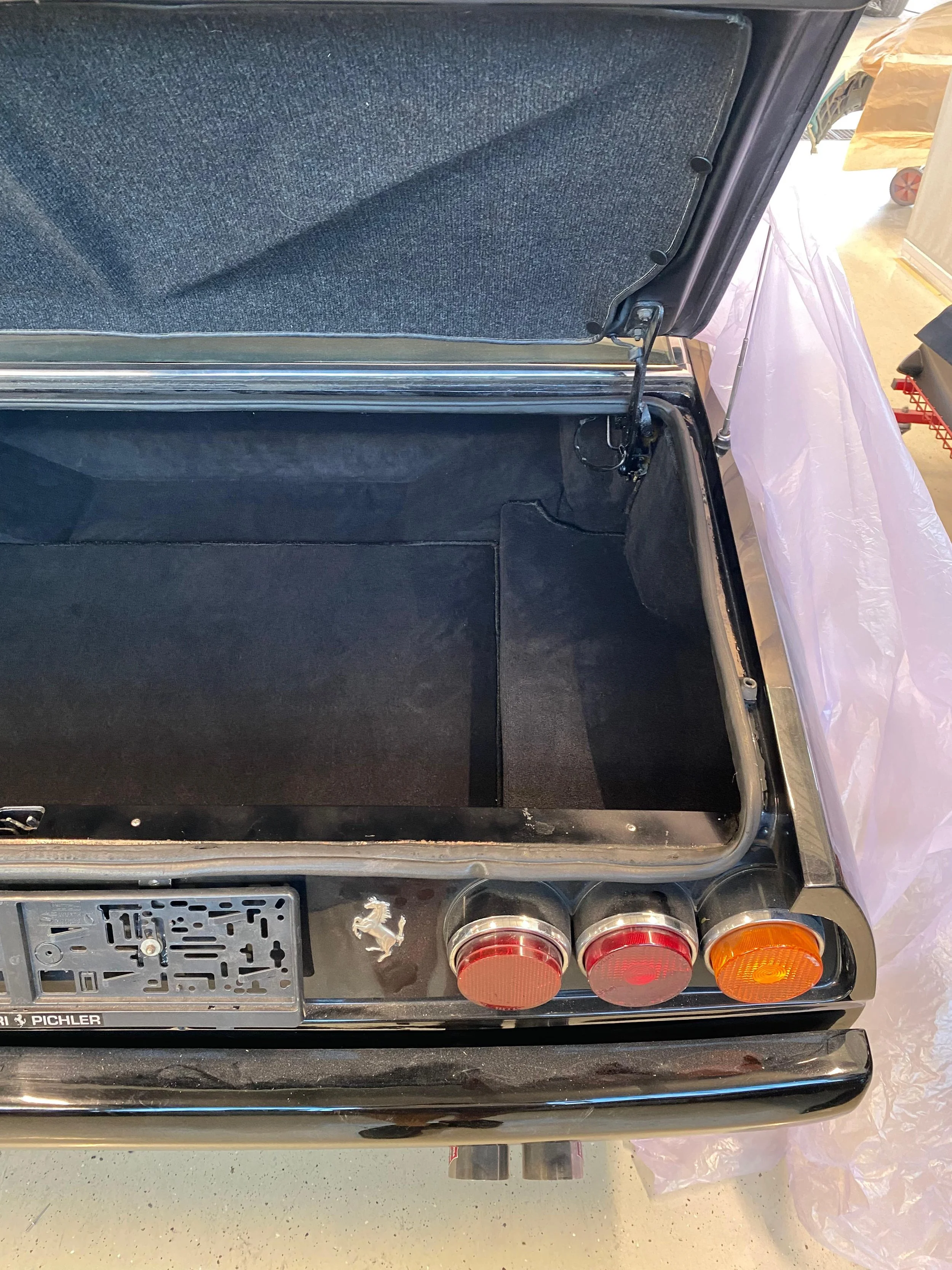 Der Kofferraum eines schwarzen Autos mit dem Ferrari-Logo, ohne Gepäck. Der Kofferraumdeckel ist offen, mit einem Kopfstützen-Stahlseil und einem schwarzen Teppich ausgekleidet.